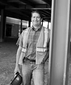 A smiling man standing outdoors near a pillar, wearing a safety vest, plaid shirt, and jeans, holding a helmet in his hand.