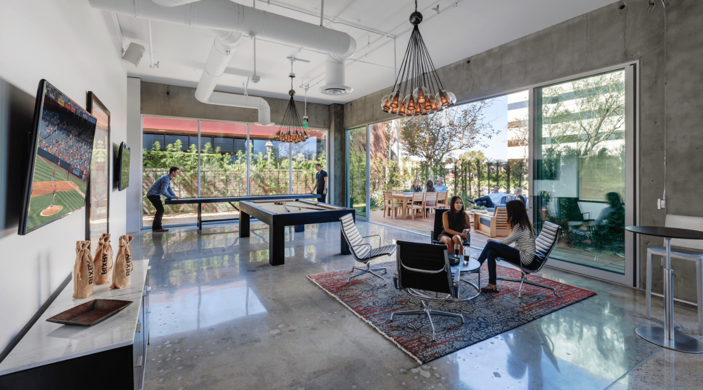 Modern lounge area with large glass windows, showing a patio with people outside, a pool table, wall-mounted TVs, and two women sitting on chairs having a conversation.