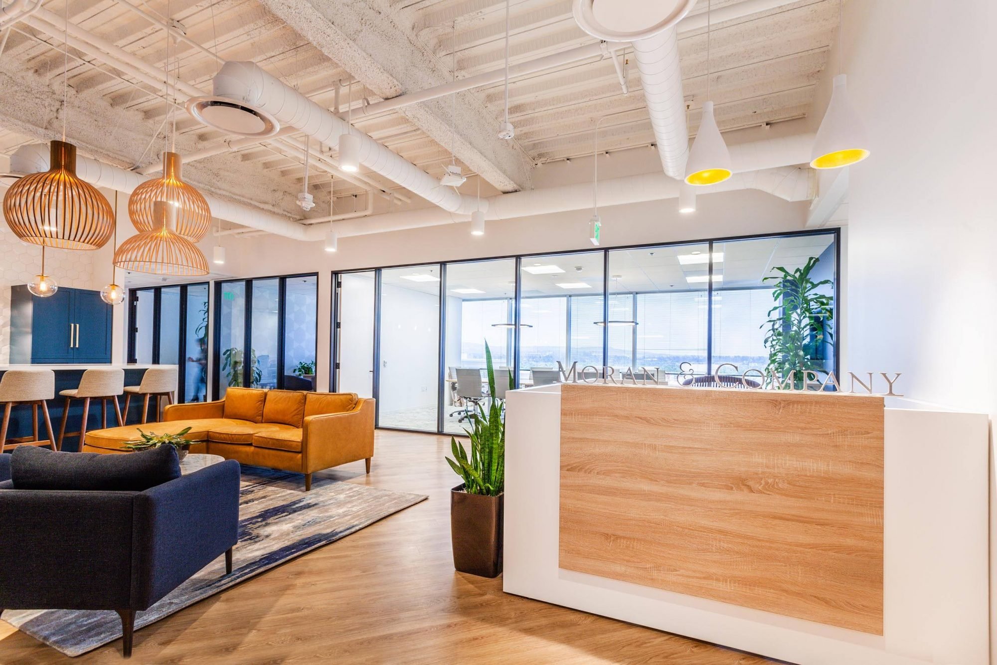 Modern office lobby with wooden reception desk, comfortable black and brown sofas, potted plants, glass walls, and decorative pendant lights in gold. Exposed ceiling with white ductwork.