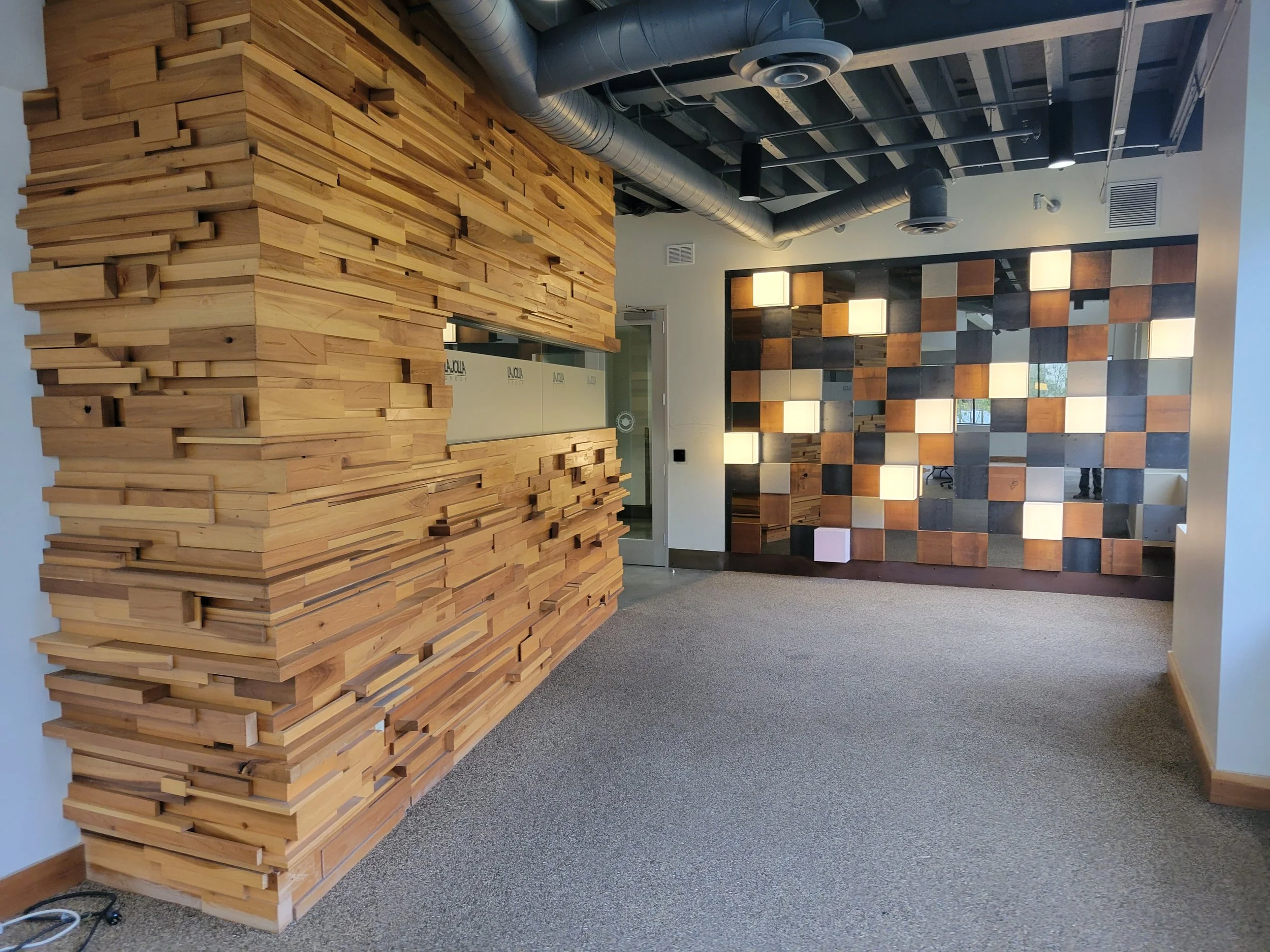 Interior of a modern office with a wooden accent wall and a partition made of checkered wooden blocks, ceiling with exposed ductwork, and carpeted floor.