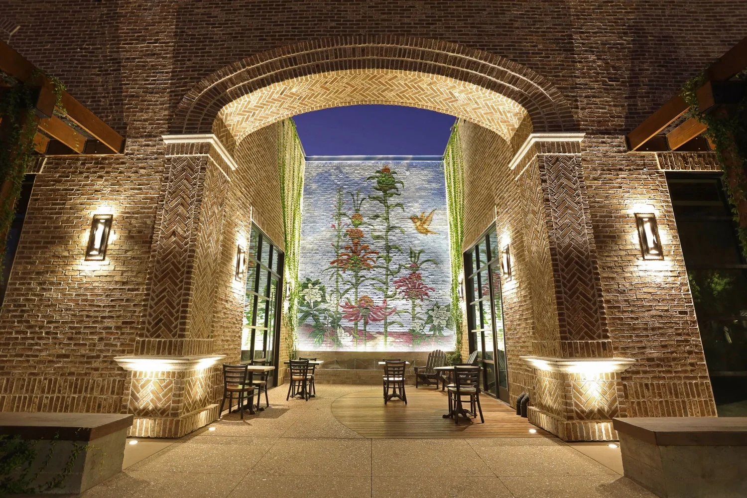 Night view of an outdoor seating area with brick arches and wall featuring a glass mural of flowers and a hummingbird, illuminated by lights.