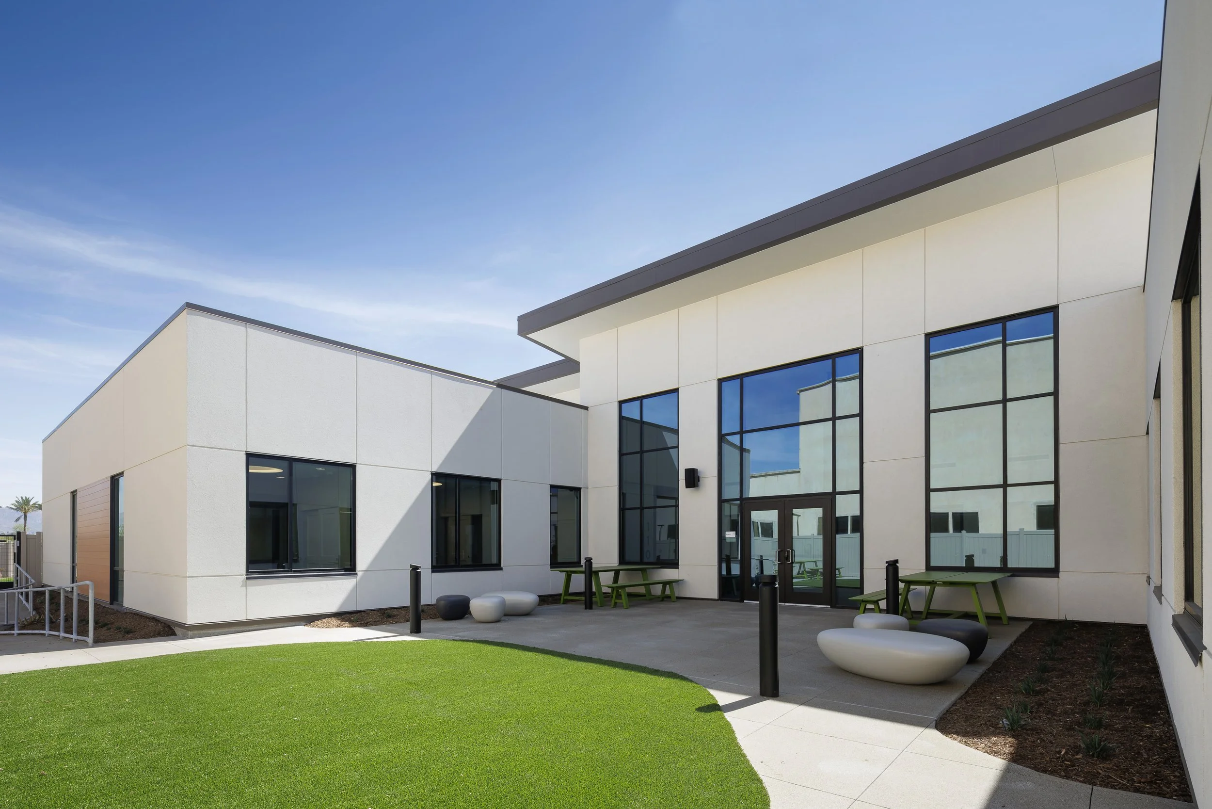 Modern white building with large glass windows, outdoor seating, and a green lawn.