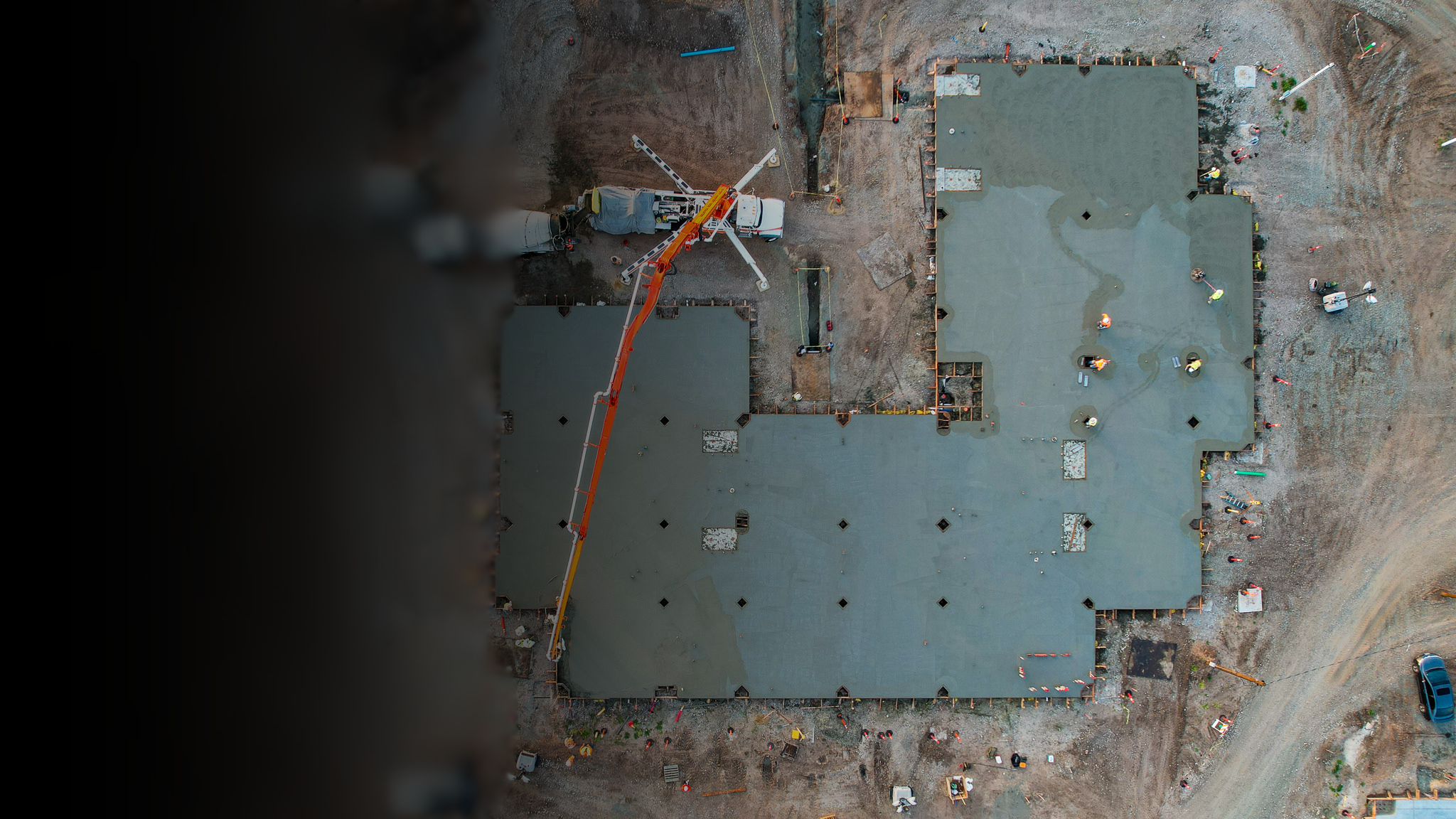 An aerial view of a construction site showing workers pouring concrete on two sections of a building foundation, with a concrete pump truck and construction materials around the area.