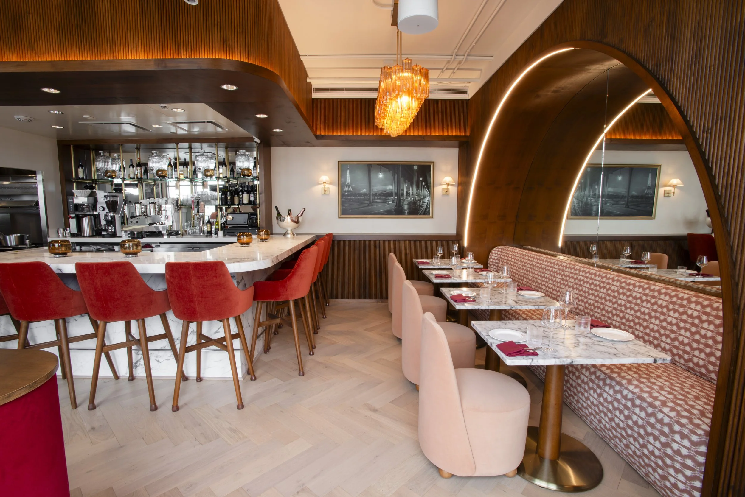 Interior of a restaurant with red cushioned chairs around marble tables, wood-paneled walls, and a bar area with glassware and bottles. There are framed black-and-white photos and warm lighting fixtures.