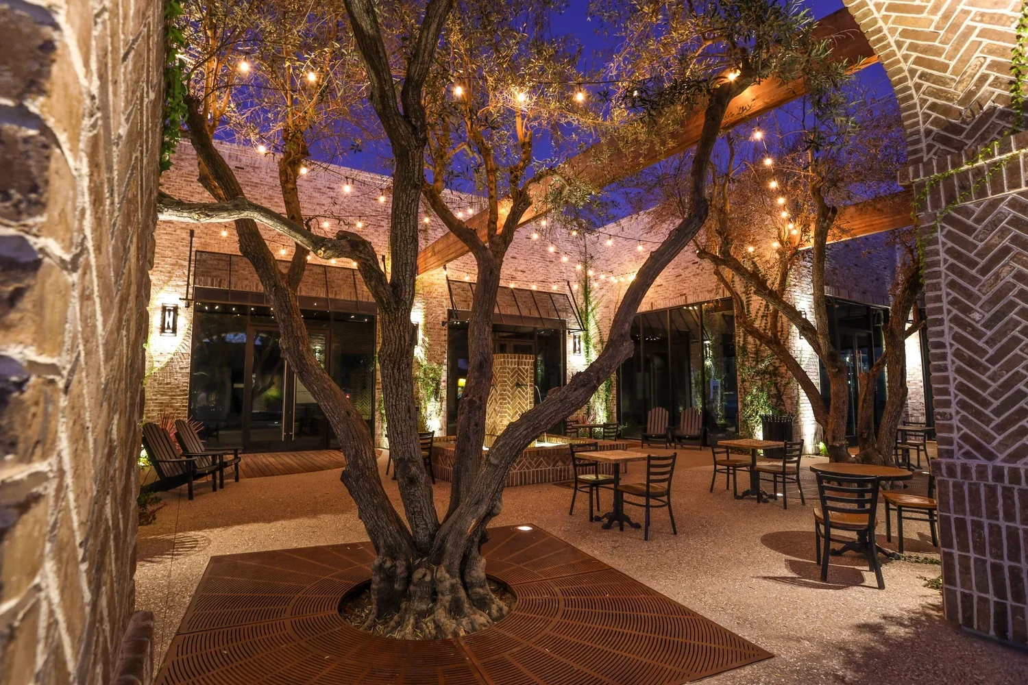 An outdoor patio area with a large, central tree surrounded by patio furniture and string lights, enclosed by brick walls and glass doors.
