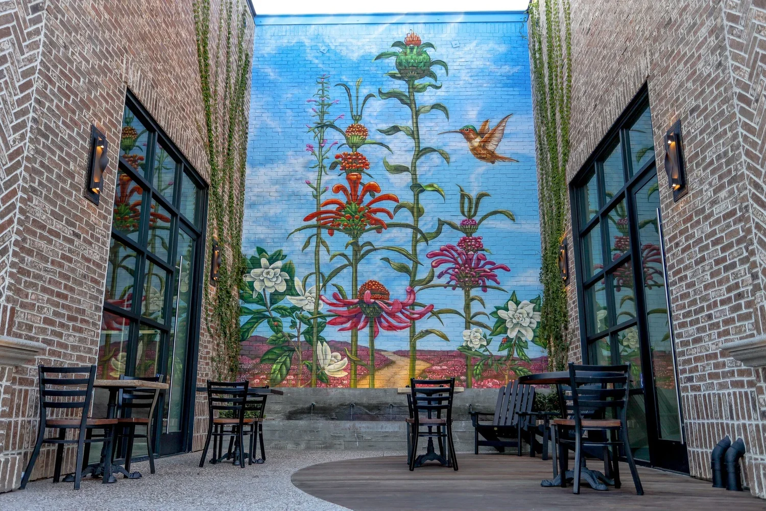 Outdoor patio with brick walls, black chairs and tables, and a large mural on the back wall depicting a garden of tall flowers and a hummingbird against a blue sky.