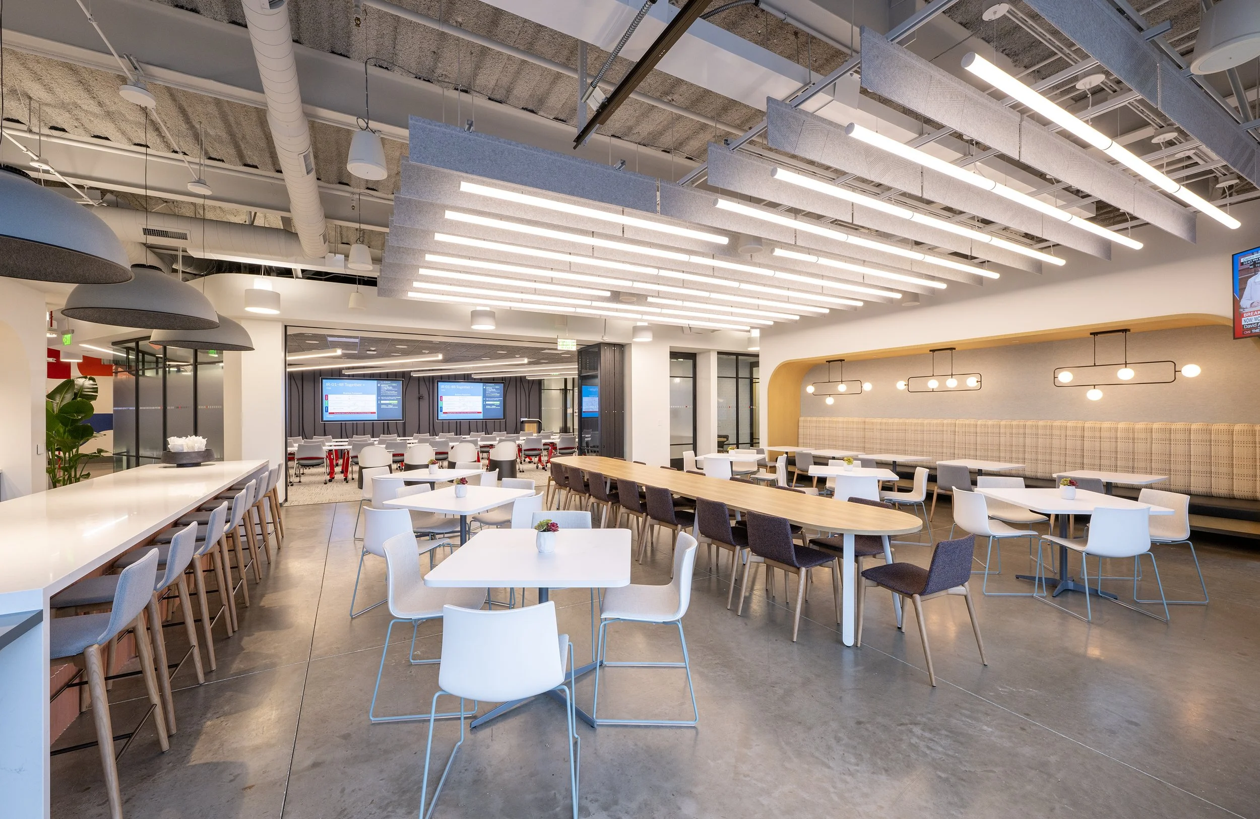 Modern cafeteria or dining area with various tables and chairs, large ceiling lights, and digital screens on the wall.