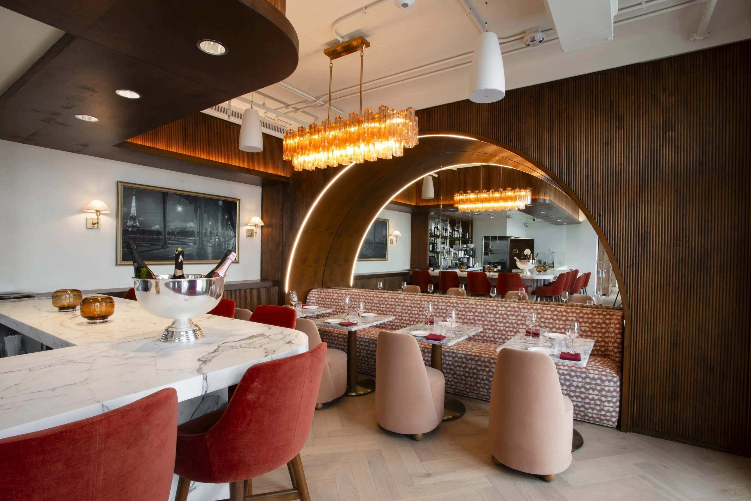A modern restaurant interior with a white marble table, red velvet chairs, and a bar area in the background. The ceiling features a large rectangular orange glass chandelier and white pendant lights. Wooden wall paneling and art pieces decorate the s