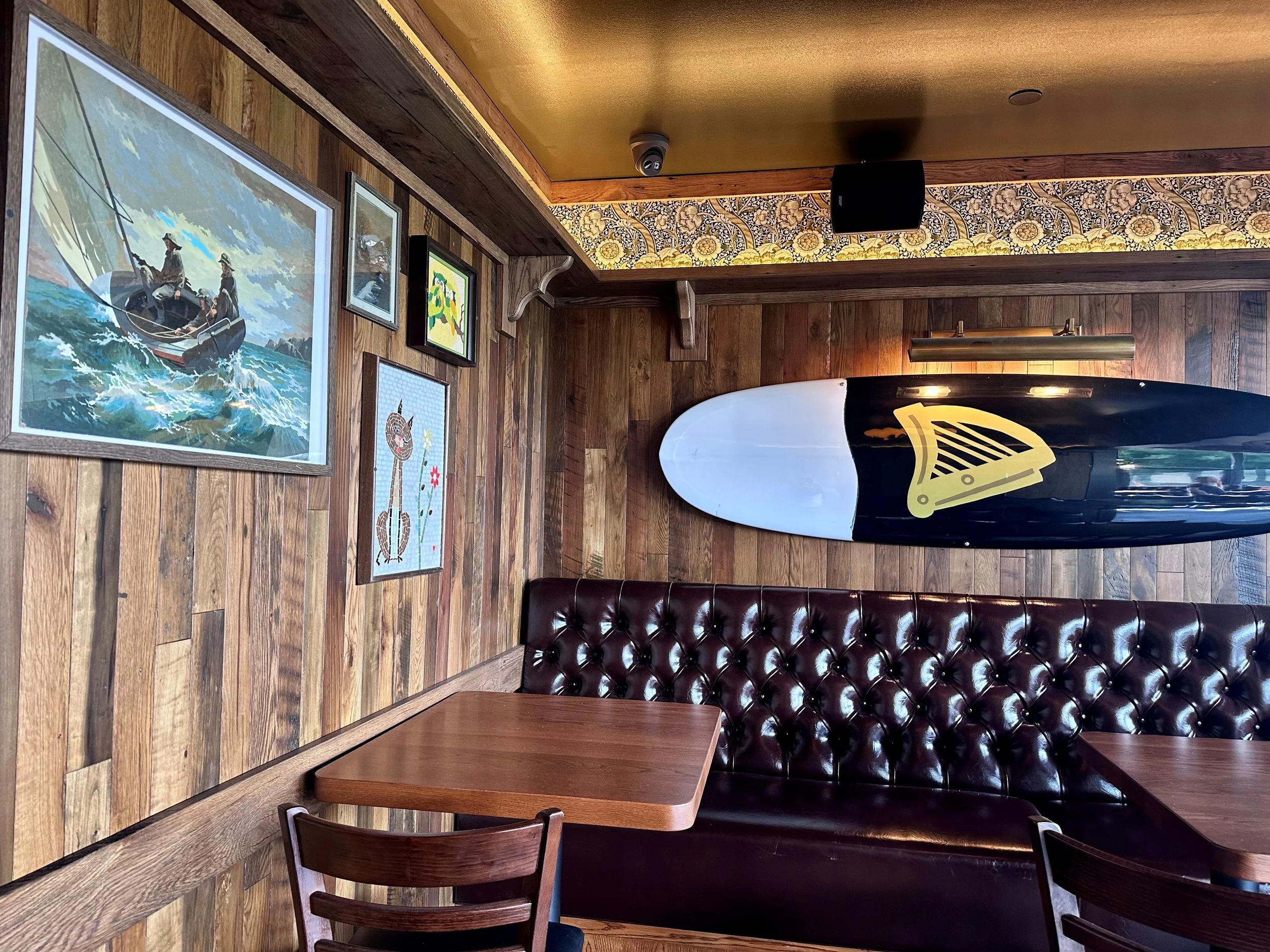 Wood-paneled interior of a restaurant or cafe with framed artwork on the wall, including a sailing scene, a cartoon character, and a giraffe, along with a surfboard with a yellow and black design leaning against the wall, and wood tables and chairs.