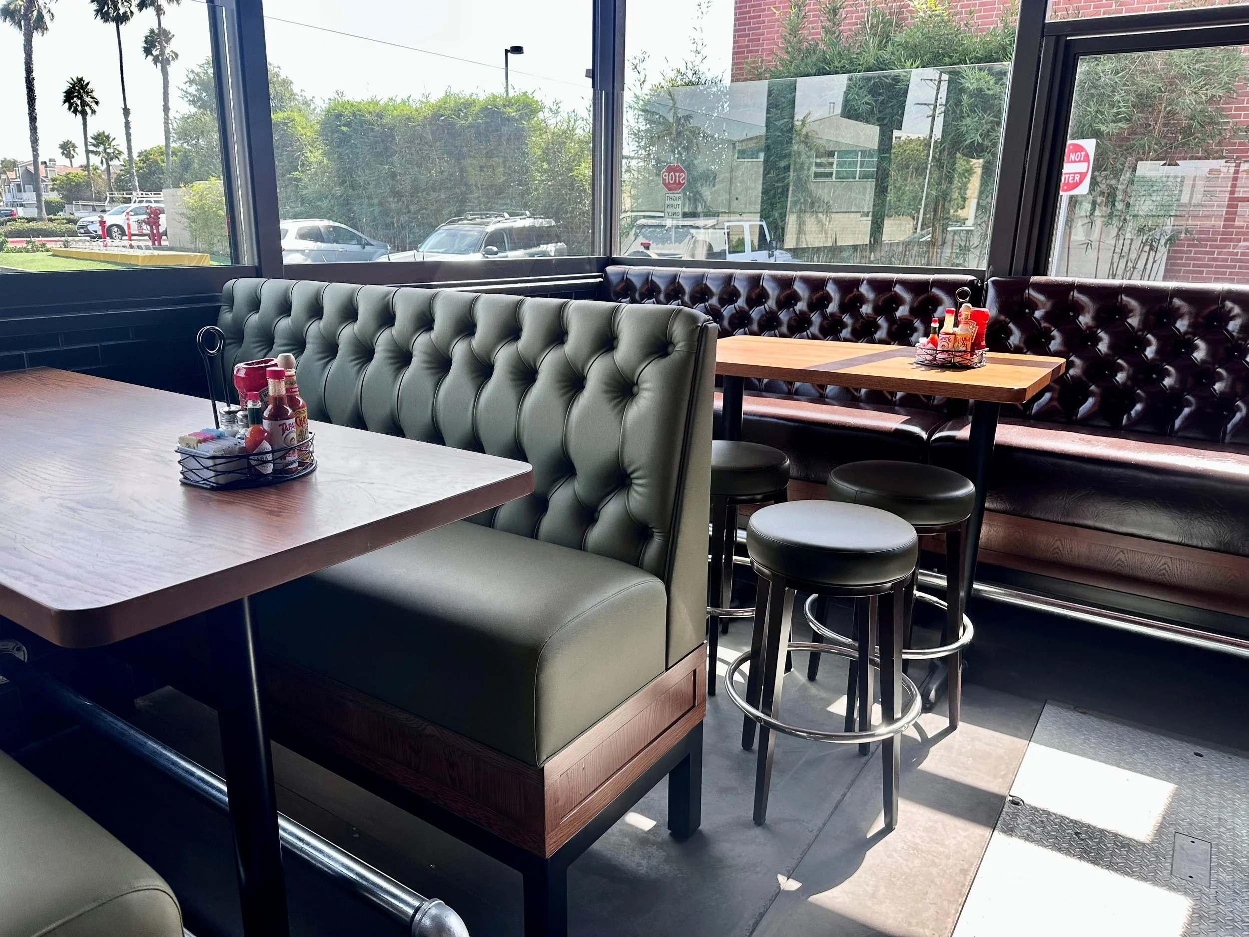 A dining area inside a restaurant with booths and tables. The booths have tufted leather upholstery, one in light green and one in dark brown. Condiment bottles and napkins are on the tables. Large windows reveal trees, parked cars, street signs, and