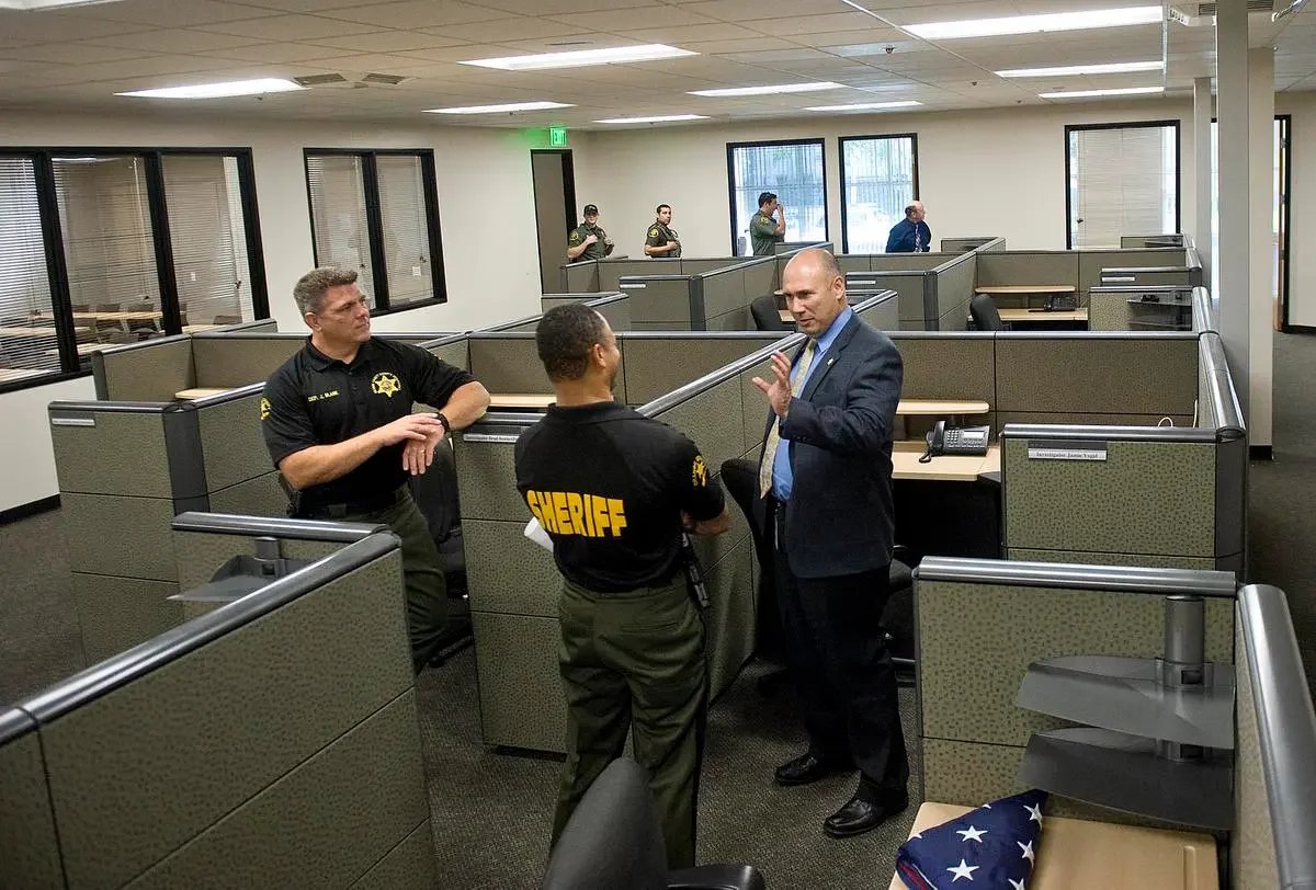 Three law enforcement officers and a man in a suit engaged in a conversation inside an office with cubicles. Four additional officers are talking near windows in the background.