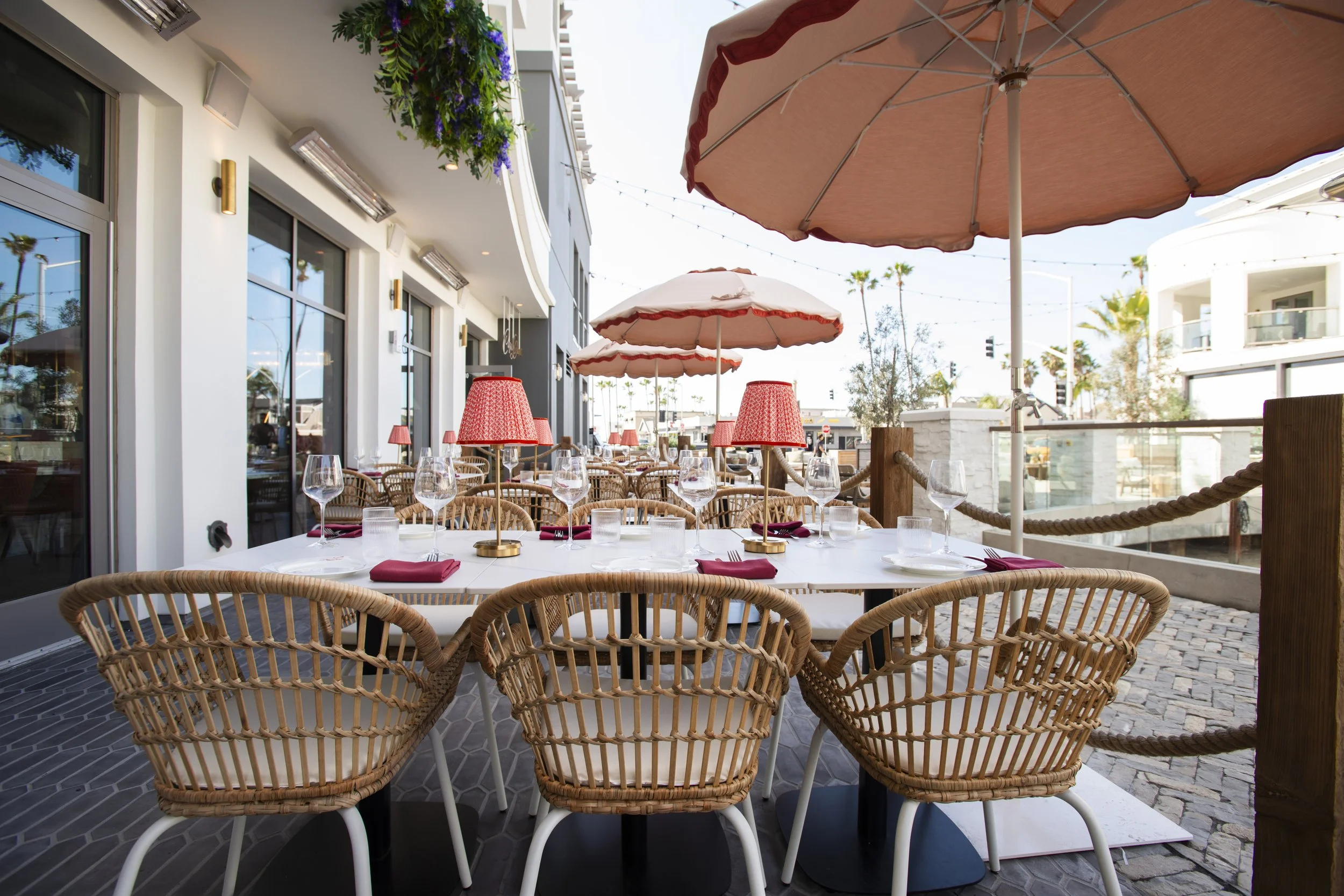 Outdoor restaurant patio with wicker chairs, glassware, and red, white, and pink lampshades on tables under large pink umbrellas, with white modern buildings and palm trees in the background.
