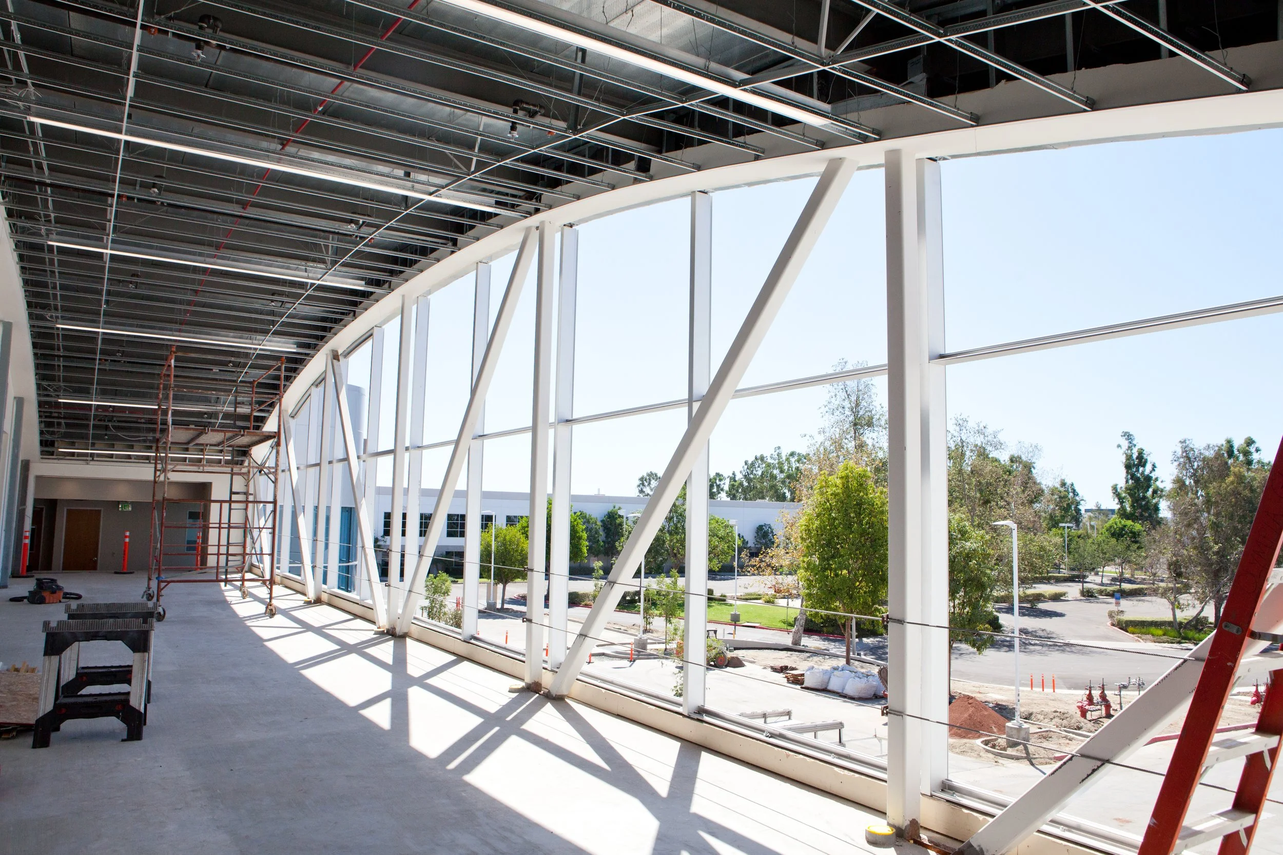 Construction site with steel framework for a building, large windows, and scaffolding inside. Outside trees and a parking lot are visible.