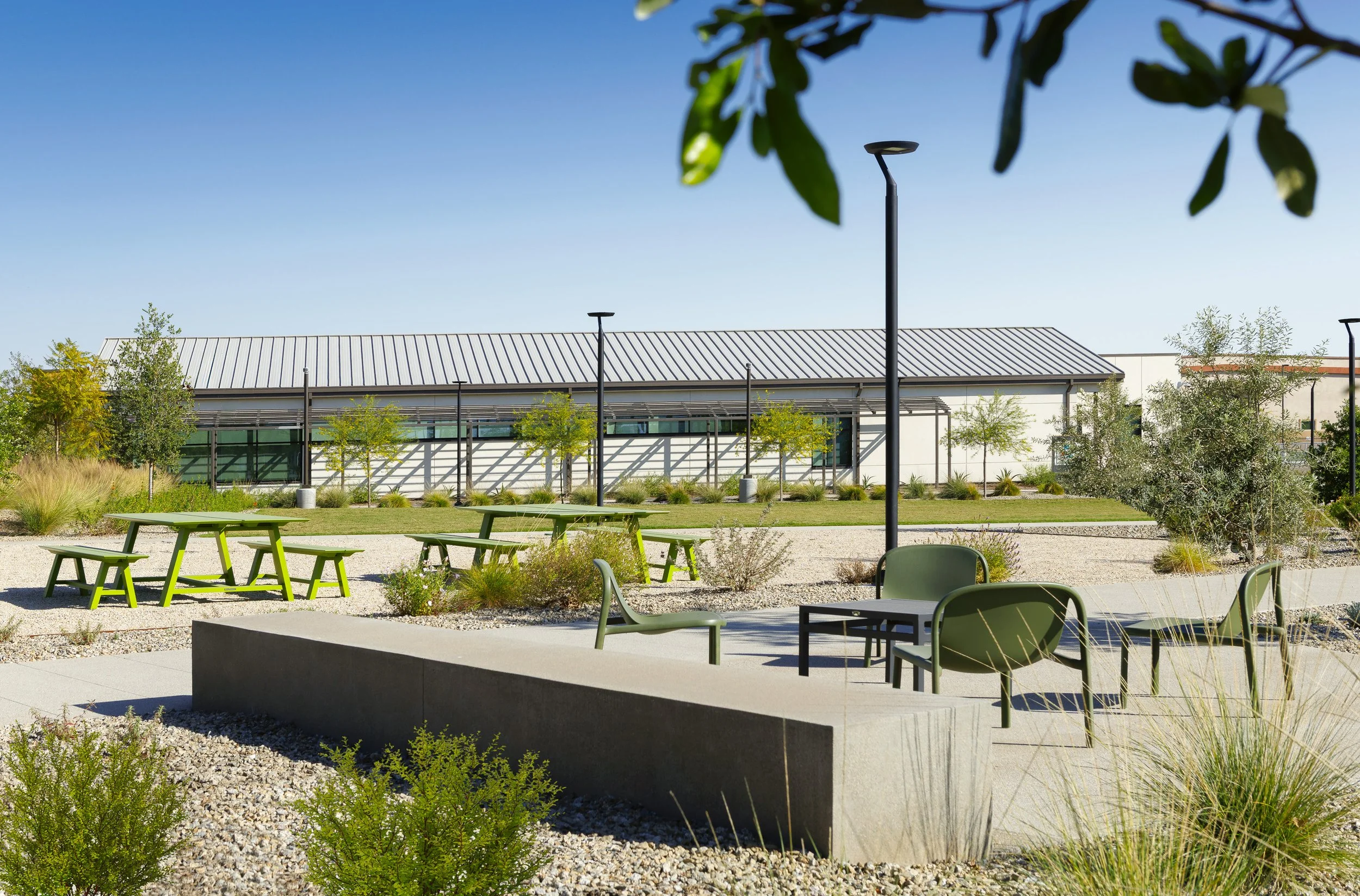 An outdoor area with green tables and chairs, landscaped with small trees, bushes, and gravel, with a modern building in the background and a clear blue sky.