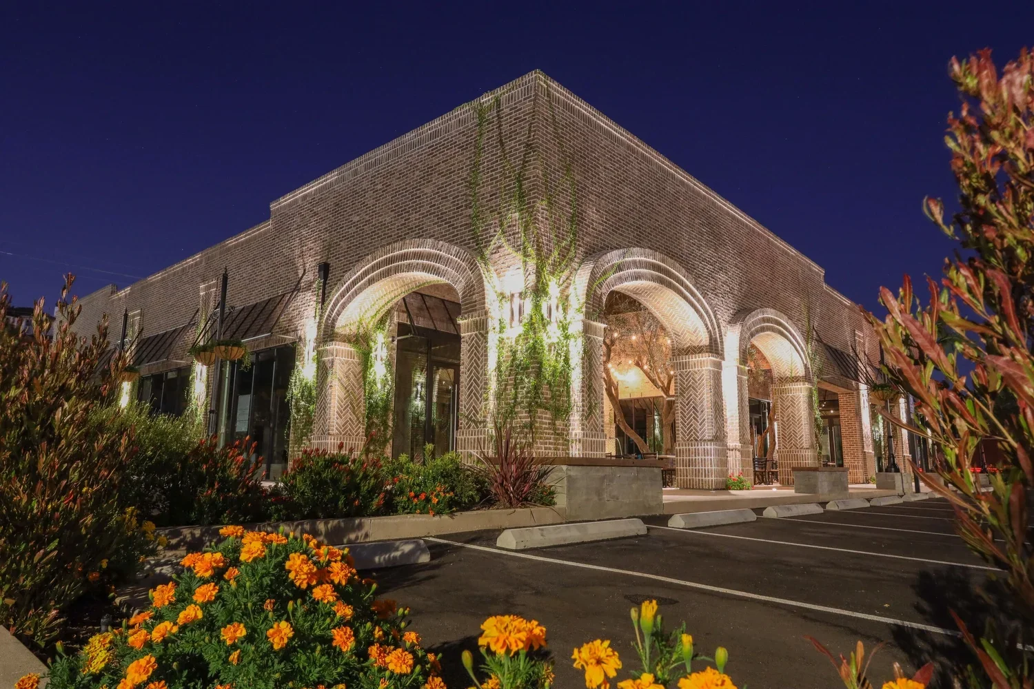 Nighttime view of a brick building with arched entrances, illuminated lights, and greenery climbing on the walls, surrounded by flowerbeds with orange and red flowers.