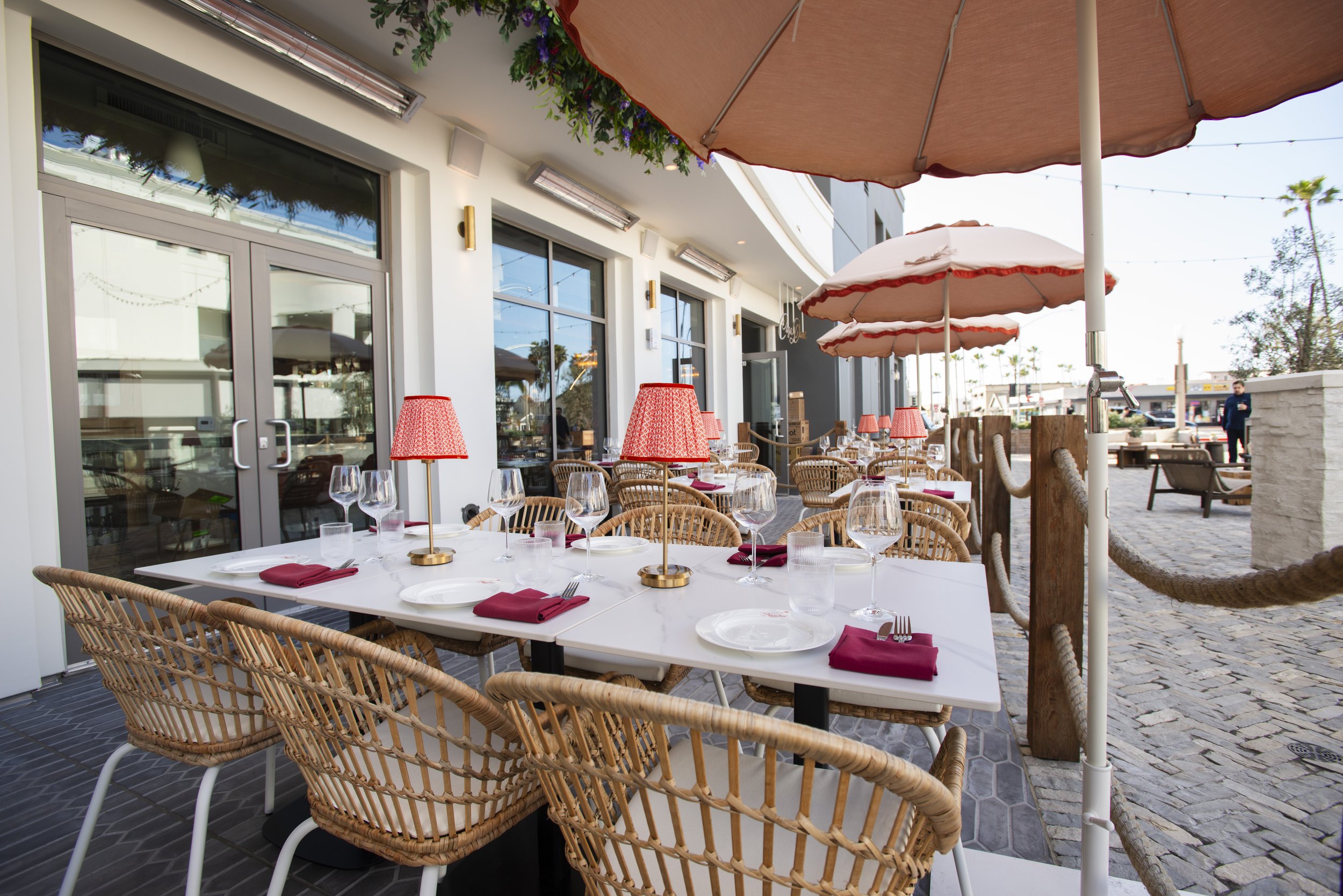 An outdoor restaurant patio with a white table set with wine glasses, plates, silverware, and red napkins, surrounded by wicker chairs, large umbrellas, and string lights.