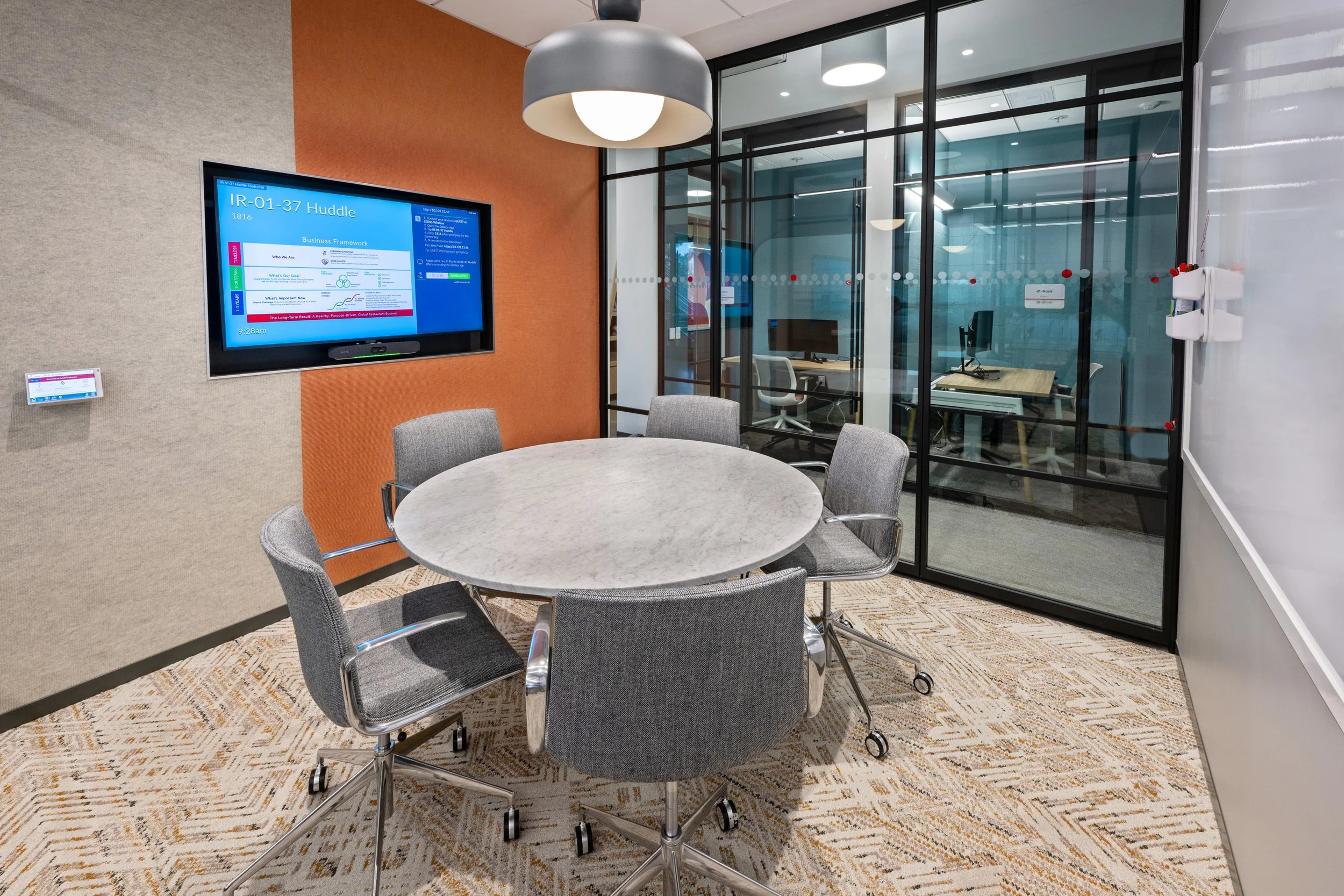 A modern office conference room with a round table, five gray chairs on wheels, a large wall-mounted digital display, and a glass-walled private office with computers and chairs inside.