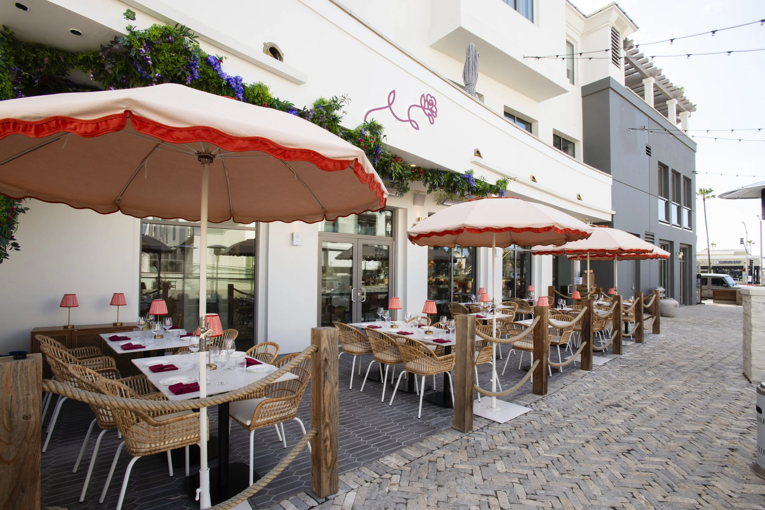 Outdoor dining area with tables set for a meal, pink umbrellas, and a modern white building with balconies and greenery, under a string of lights.