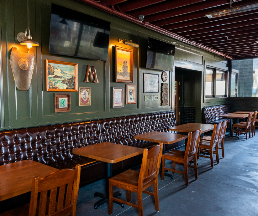 Interior view of a restaurant with dark green walls, wood tables, and wooden chairs. Decor includes framed paintings, a mounted mask, and flat-screen TVs on the wall.