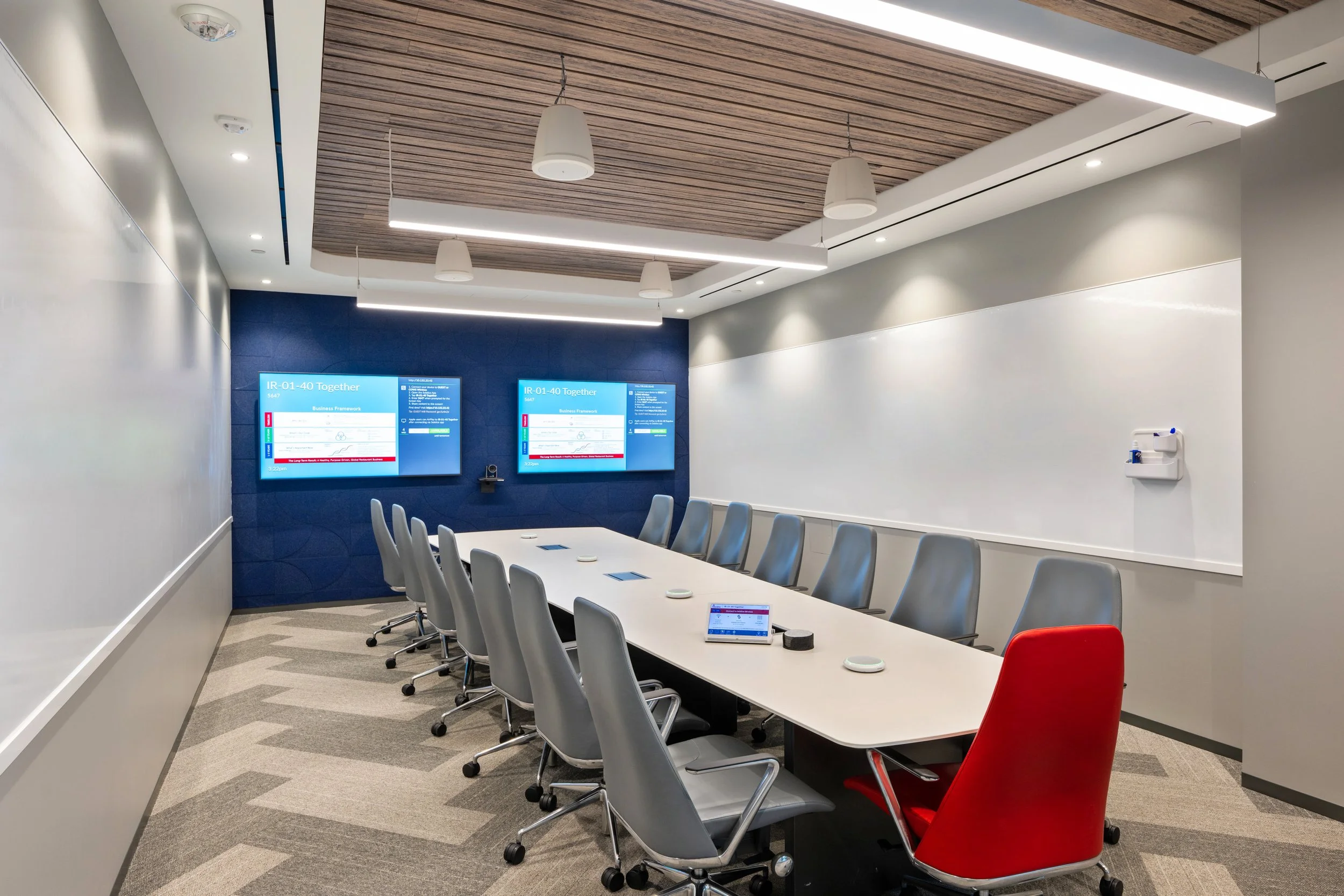 Modern conference room with a long white table surrounded by gray and red chairs. Two digital screens display a presentation on a blue wall, and a whiteboard is on the adjacent wall. The ceiling has wooden panels with hanging and recessed lighting.