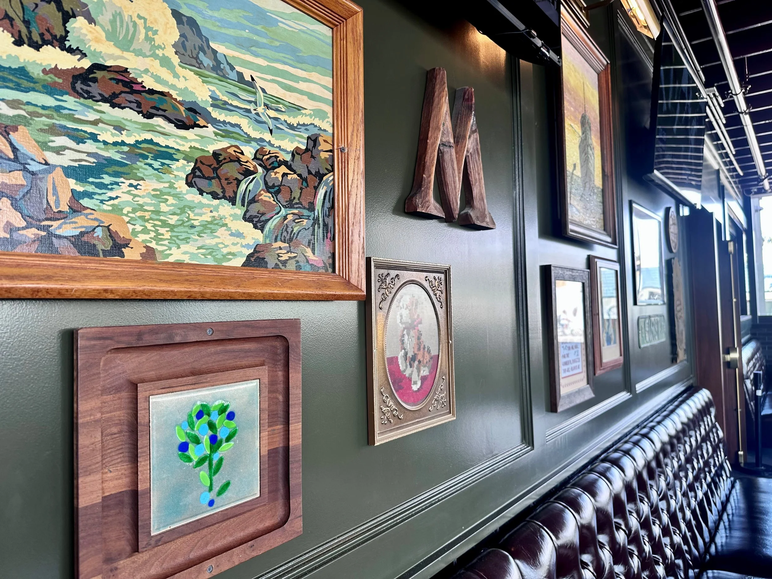 A gallery wall with framed artwork and decorations, including a picture of a river scene with rocks and waterfalls, a framed embroidery of a flower, and an abstract piece, with a row of leather chairs underneath.