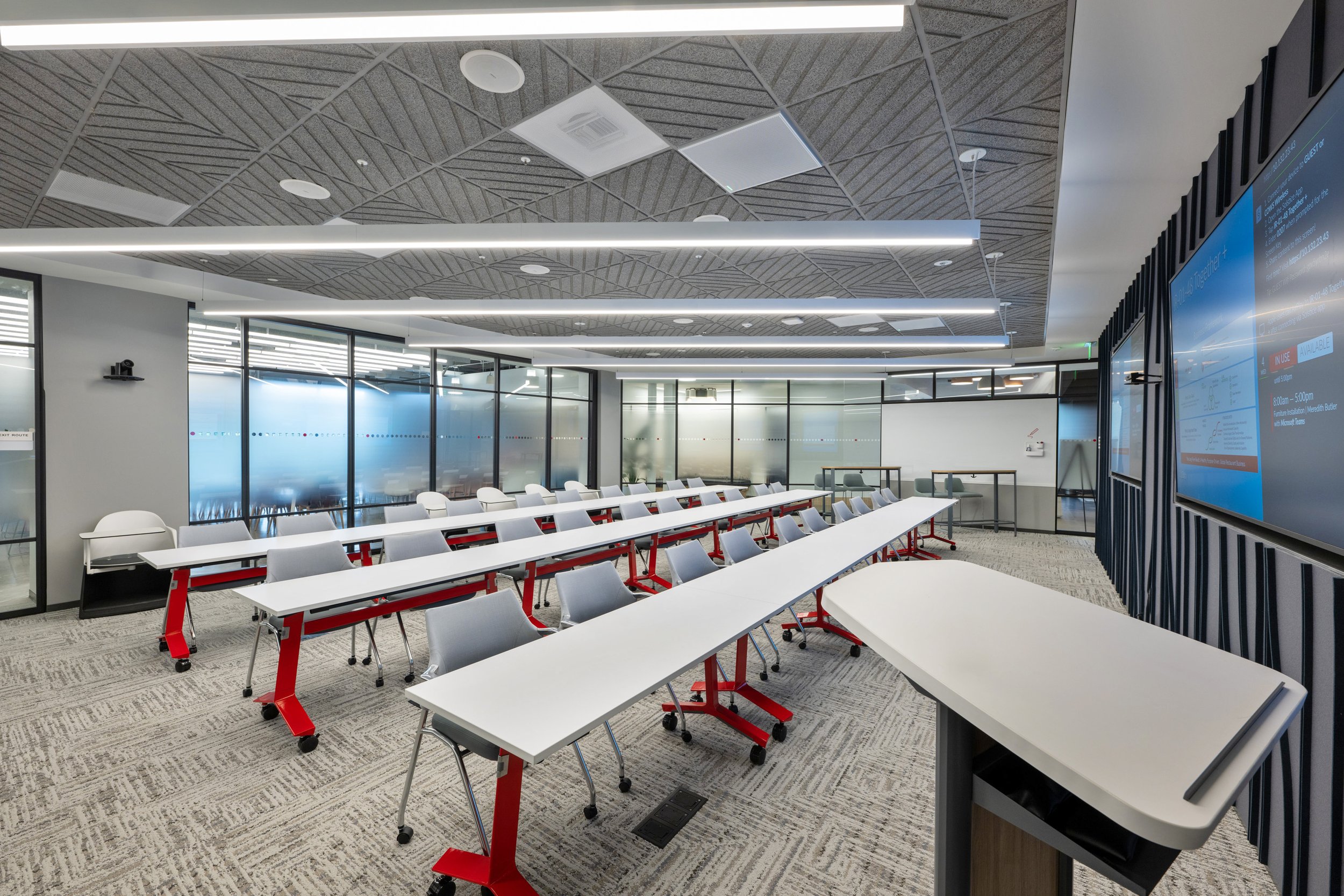 An empty modern conference room with rows of white tables and chairs on red and black wheels, large frosted glass windows, and a digital display screen on the right wall.