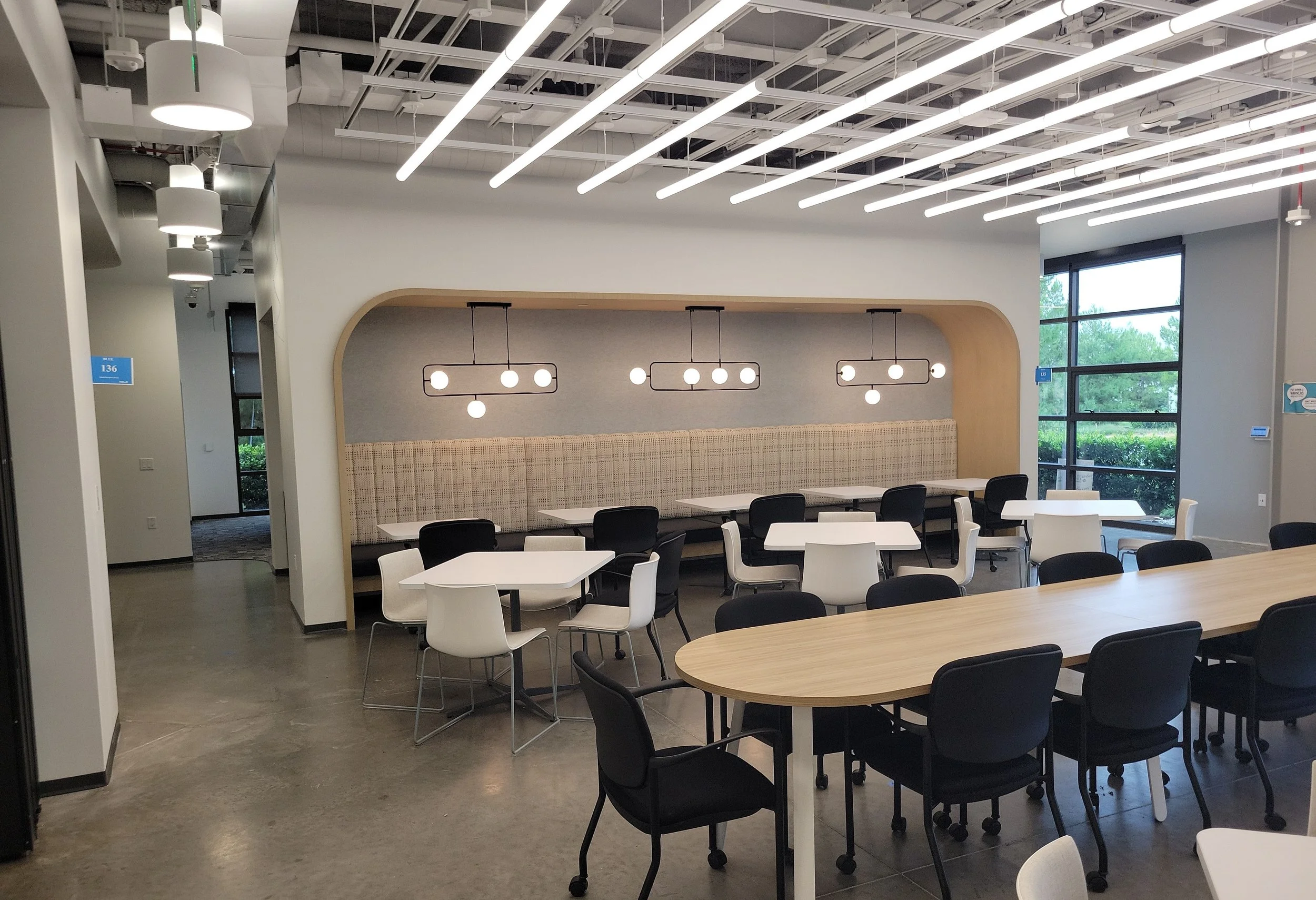 An interior of a modern, well-lit conference or dining room with contemporary lighting fixtures, large windows with greenery outside, and arranged tables and chairs.
