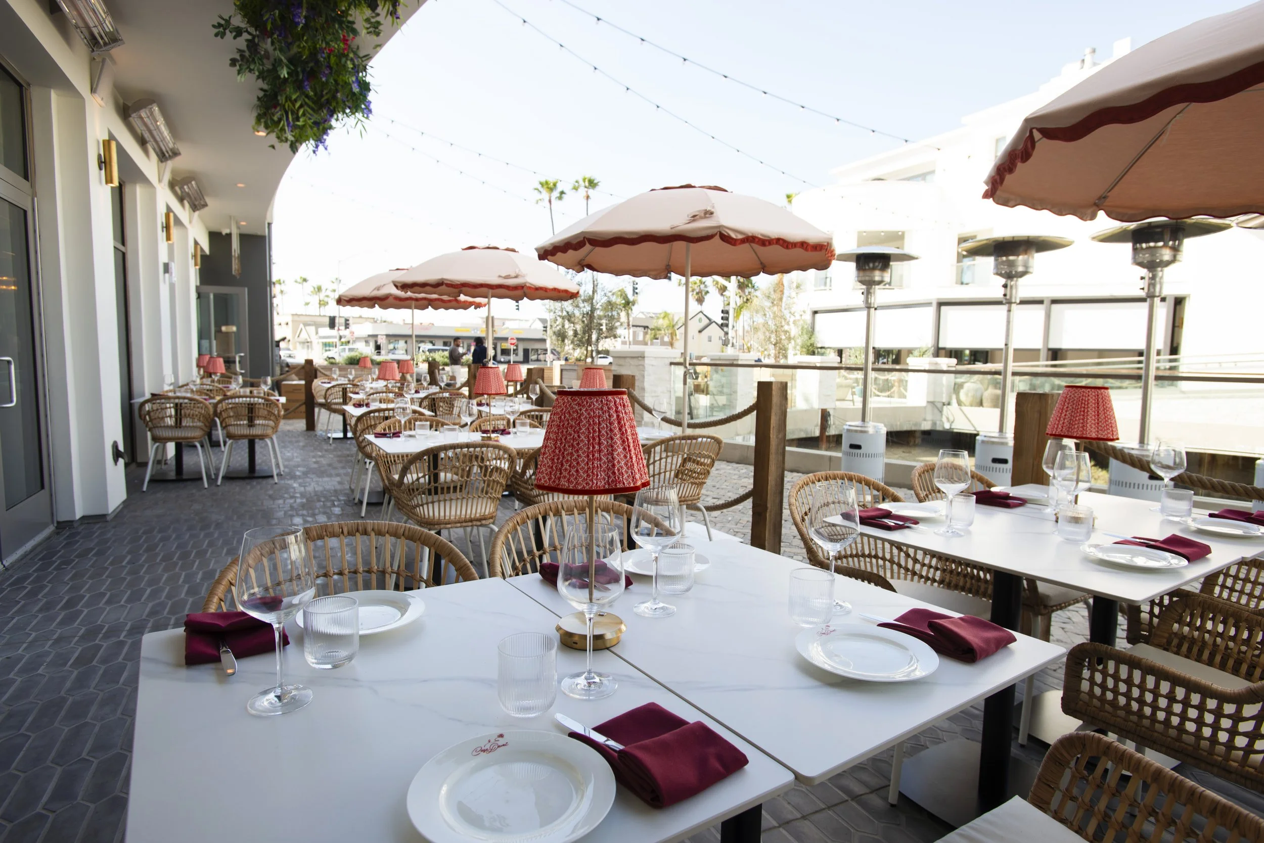 Outdoor dining area with white tables set for a meal, featuring glasses, plates, and maroon napkins. Elegant wicker chairs surround the tables. Large umbrellas provide shade, and string lights are hanging above. The setting appears to be on a patio o