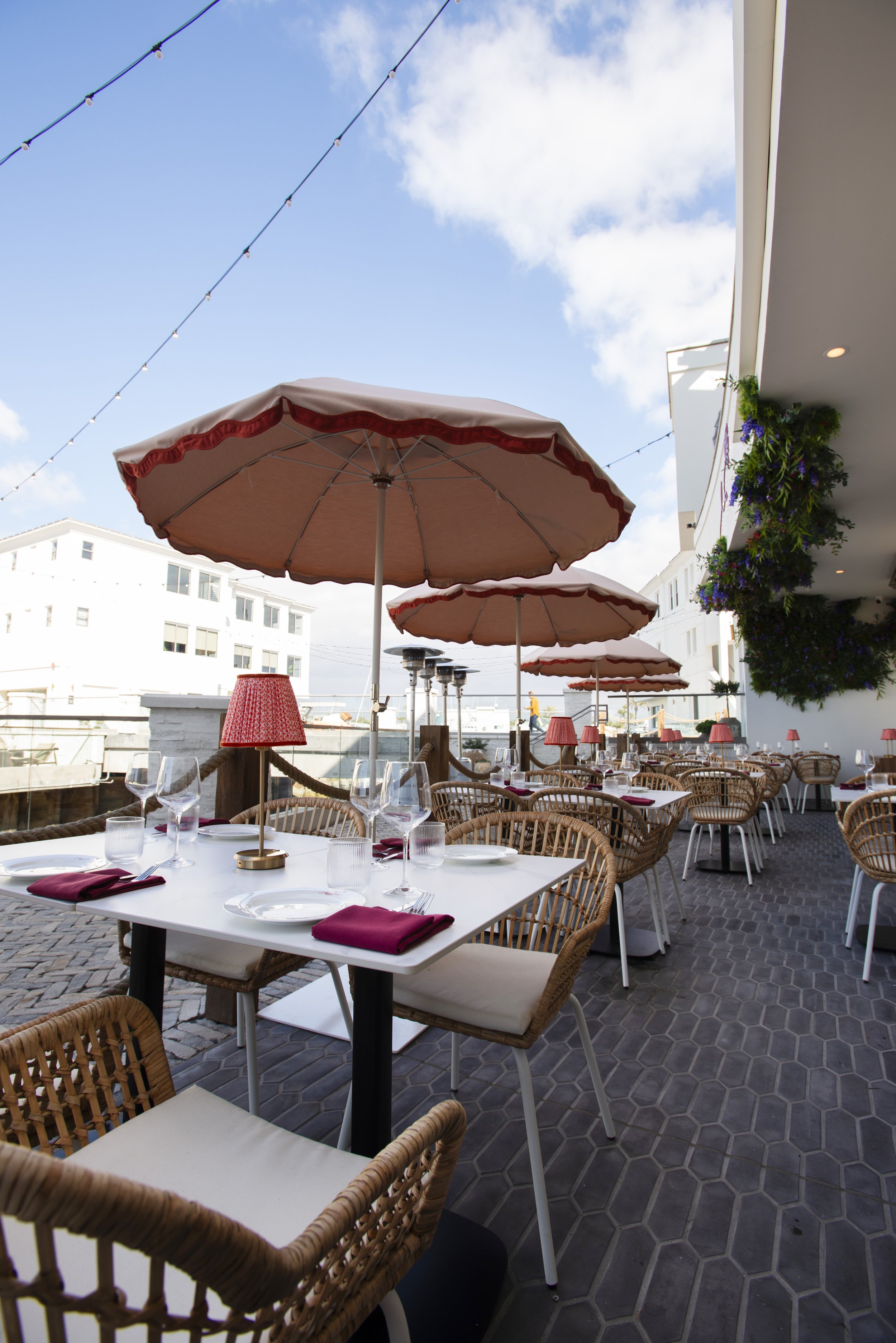 Outdoor restaurant patio with white tables set with glasses, napkins, and cutlery, surrounded by wicker chairs. Large pink umbrellas provide shade. String lights are overhead, and a building with white walls and balconies is visible in the background, along with potted purple and green plants on the balcony.