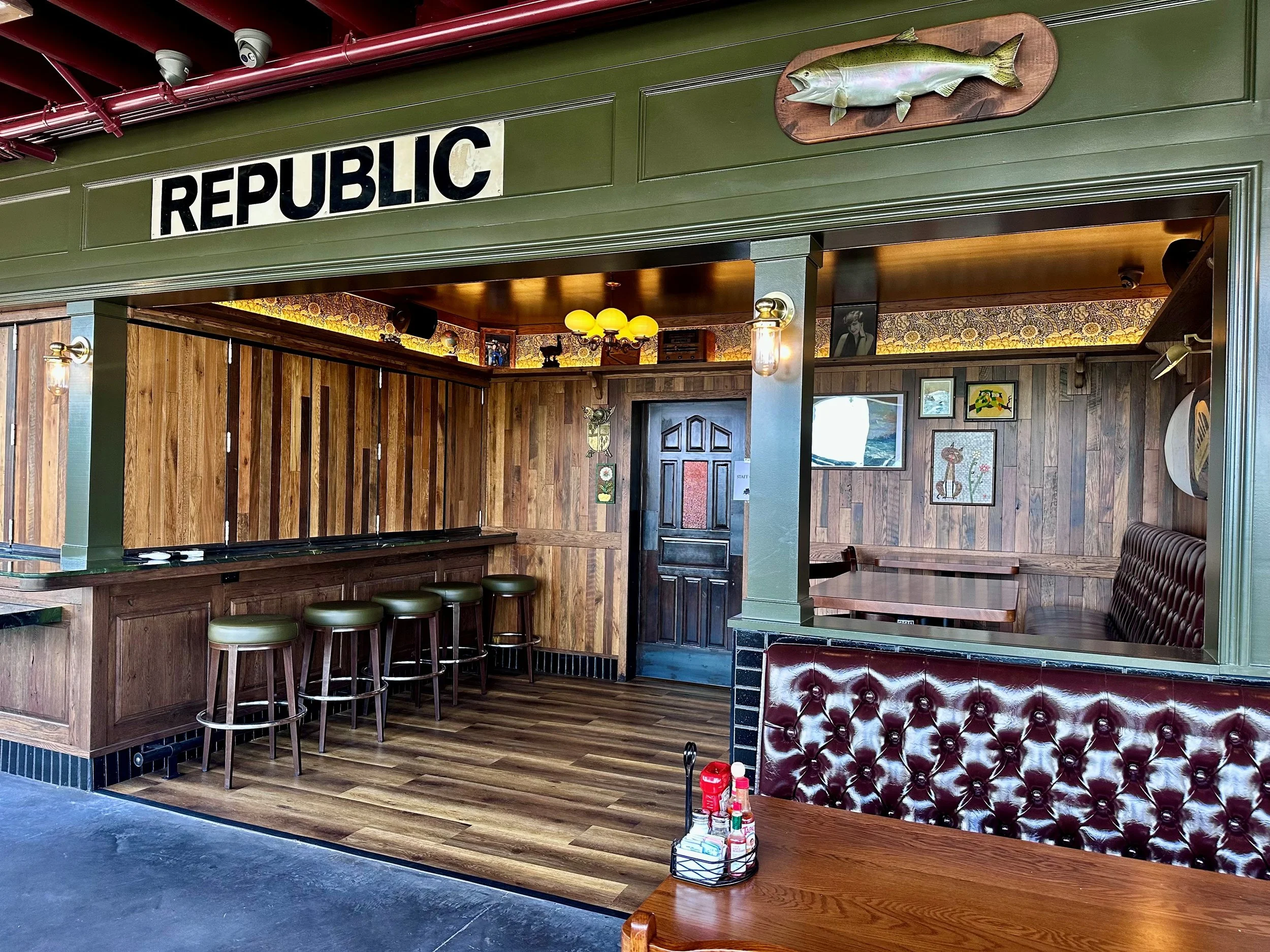 Interior of a rustic restaurant or bar with wood-paneled walls, bar stools, a black door, framed pictures, and a mounted fish on the wall. The sign 'REPUBLIC' is displayed above the entrance.