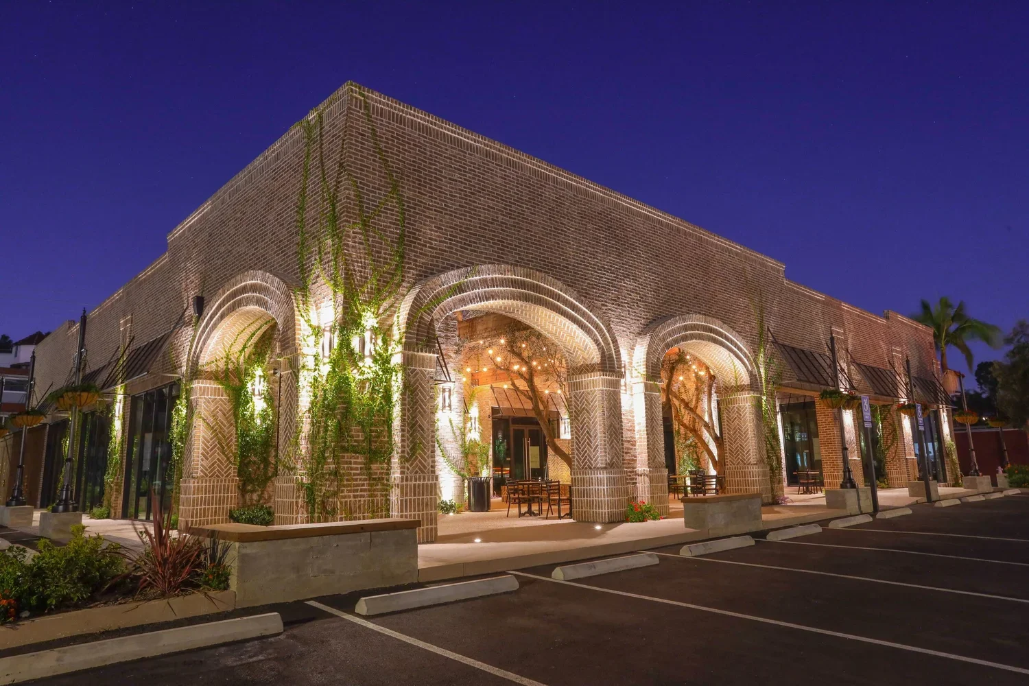 Night view of a brick commercial building with arched entryways, decorative lighting, and climbing vines, with a parking lot in foreground.