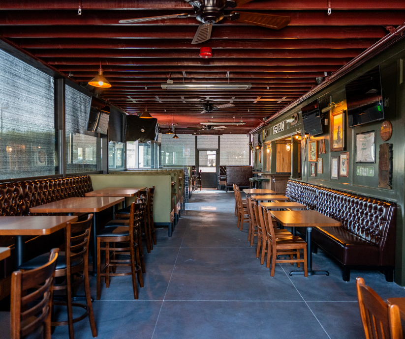 Empty restaurant with wooden tables and chairs, a green wall with framed pictures, and large windows with blinds.