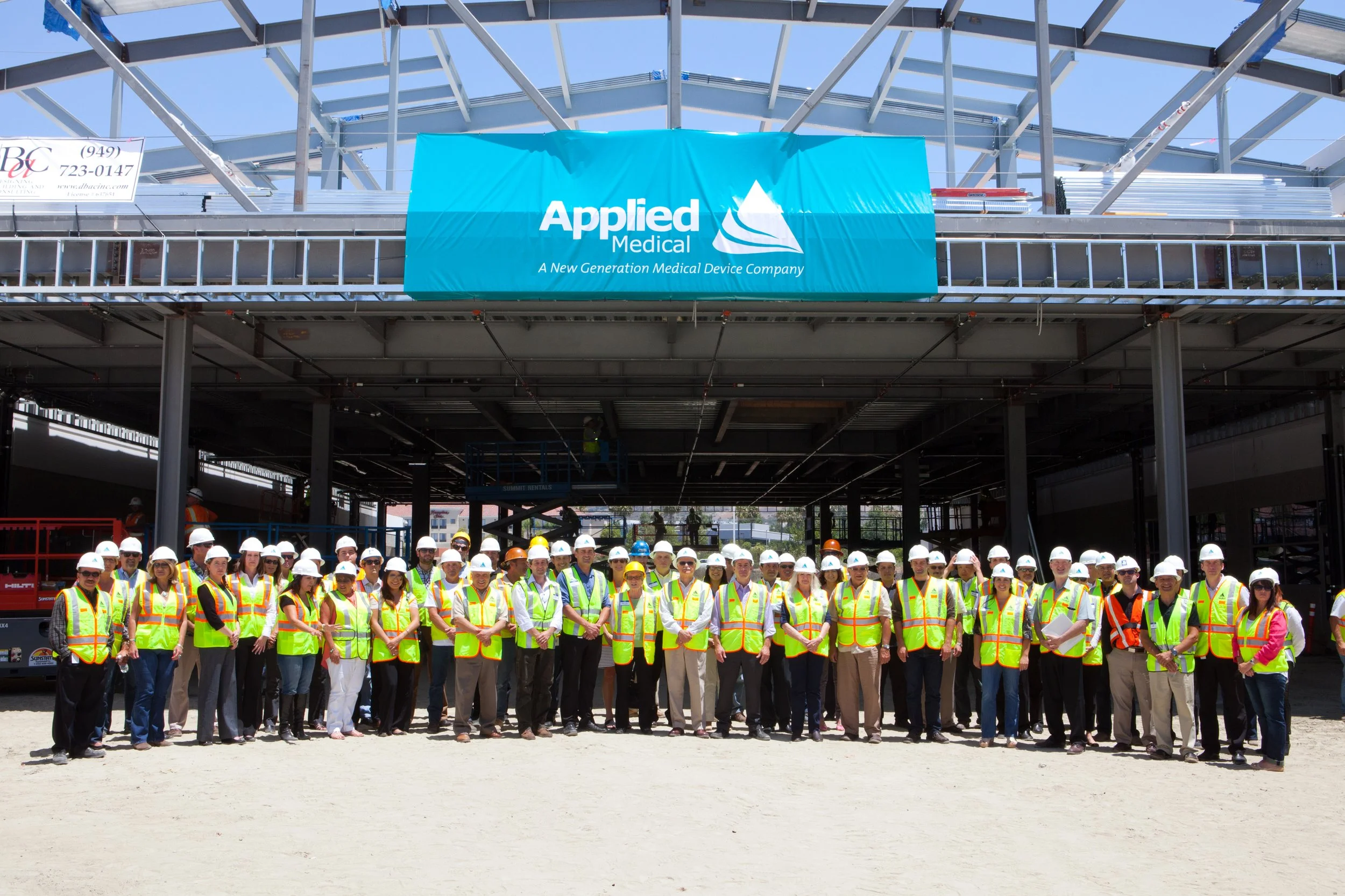 Group of construction workers and engineers wearing safety vests and helmets standing in front of a building under construction for Applied Medical, a medical device company.