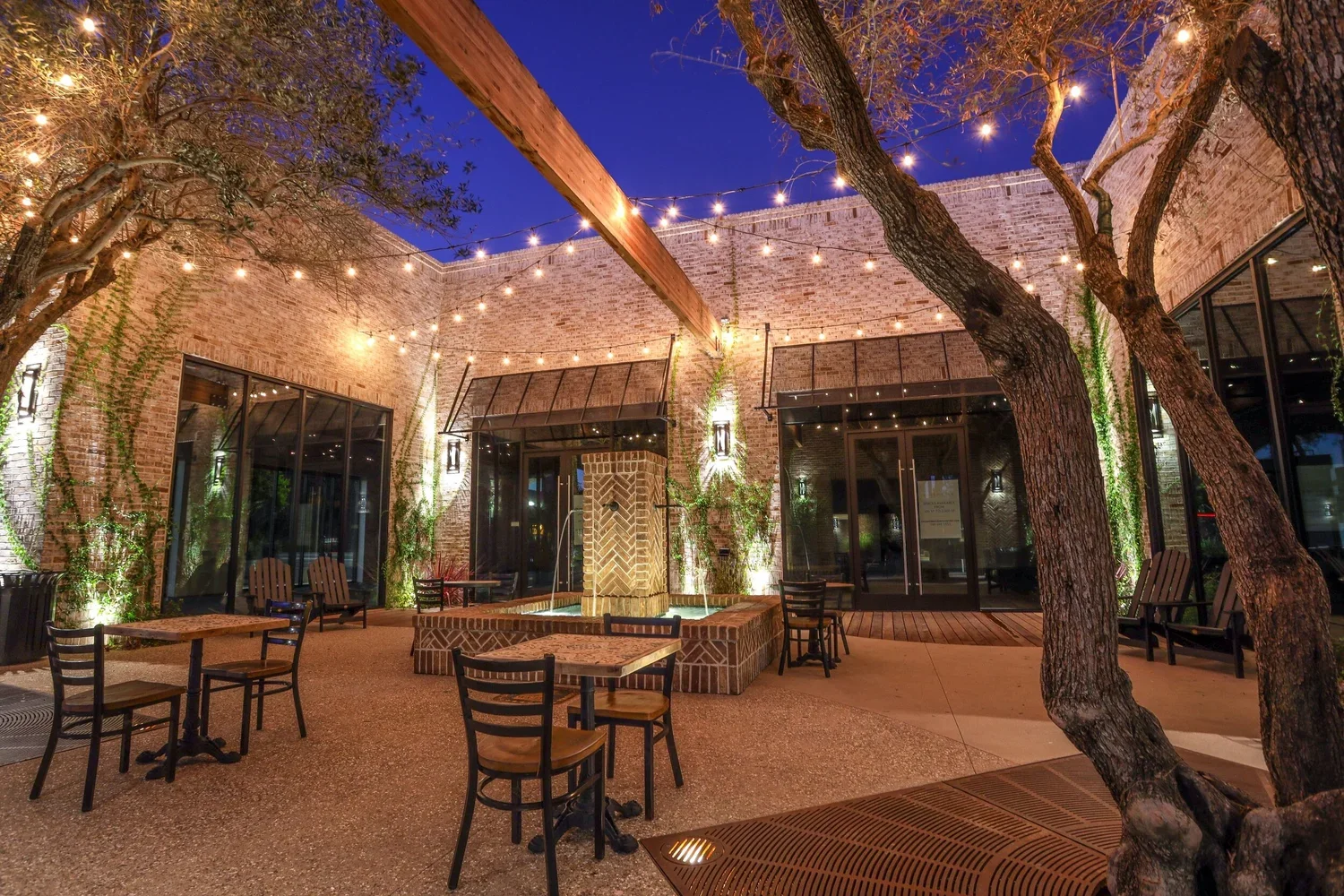 An outdoor patio area in the evening with string lights hanging overhead. There are several small tables and chairs, and a brick fountain in the center surrounded by brick ledges. The walls are brick with some greenery climbing up, and large glass do