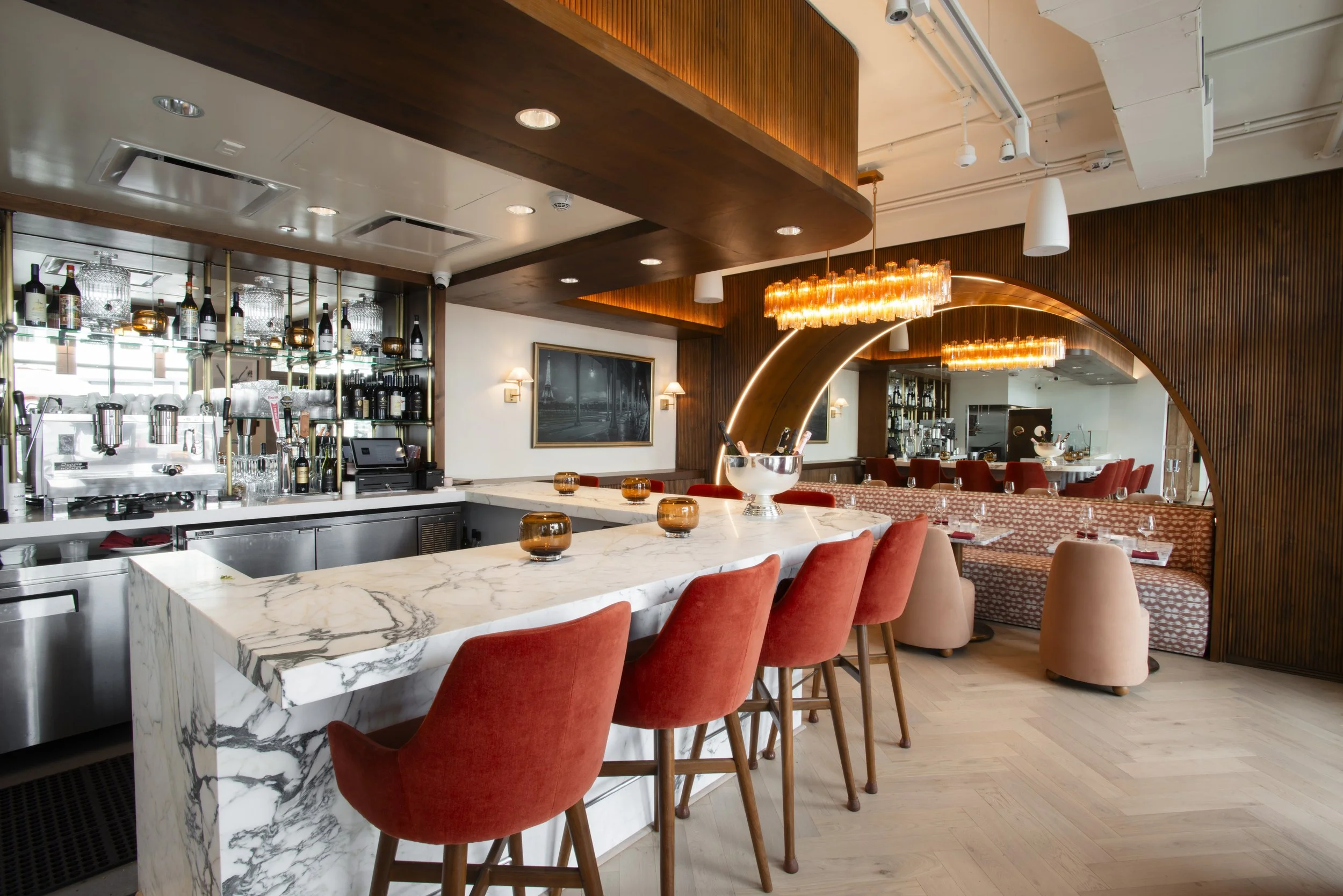 Modern restaurant interior with a marble bar counter, red upholstered chairs, wooden wall paneling, decorative lighting, and bar shelves with bottles and glasses.