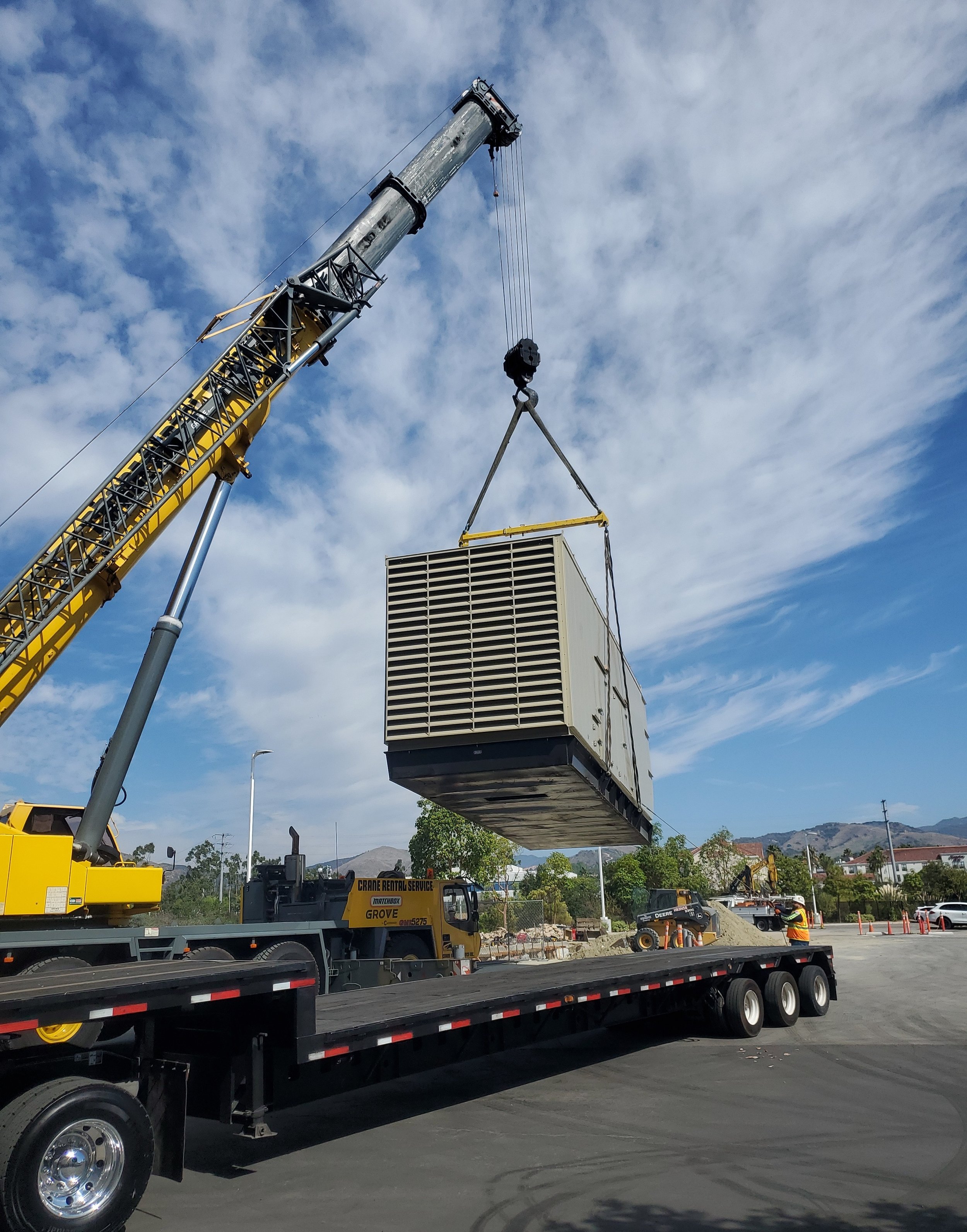 A crane lifting a large industrial air conditioning unit onto a flatbed truck at a construction site with workers and other equipment in the background.