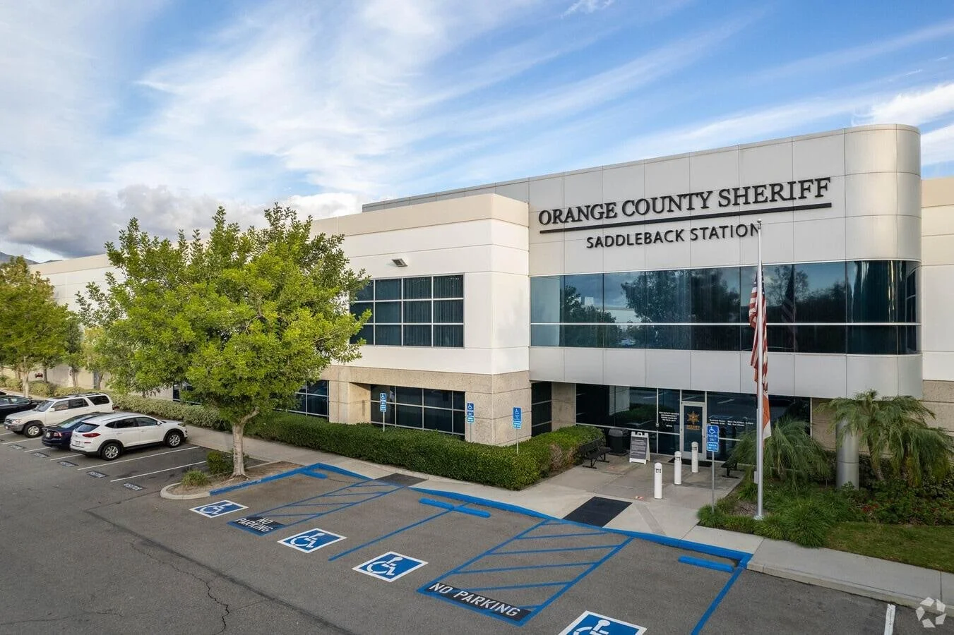 Exterior of Orange County Sheriff's Saddleback Station with a parking lot including designated handicapped parking spaces, a tree, and a flagpole with an American flag in front of the building.