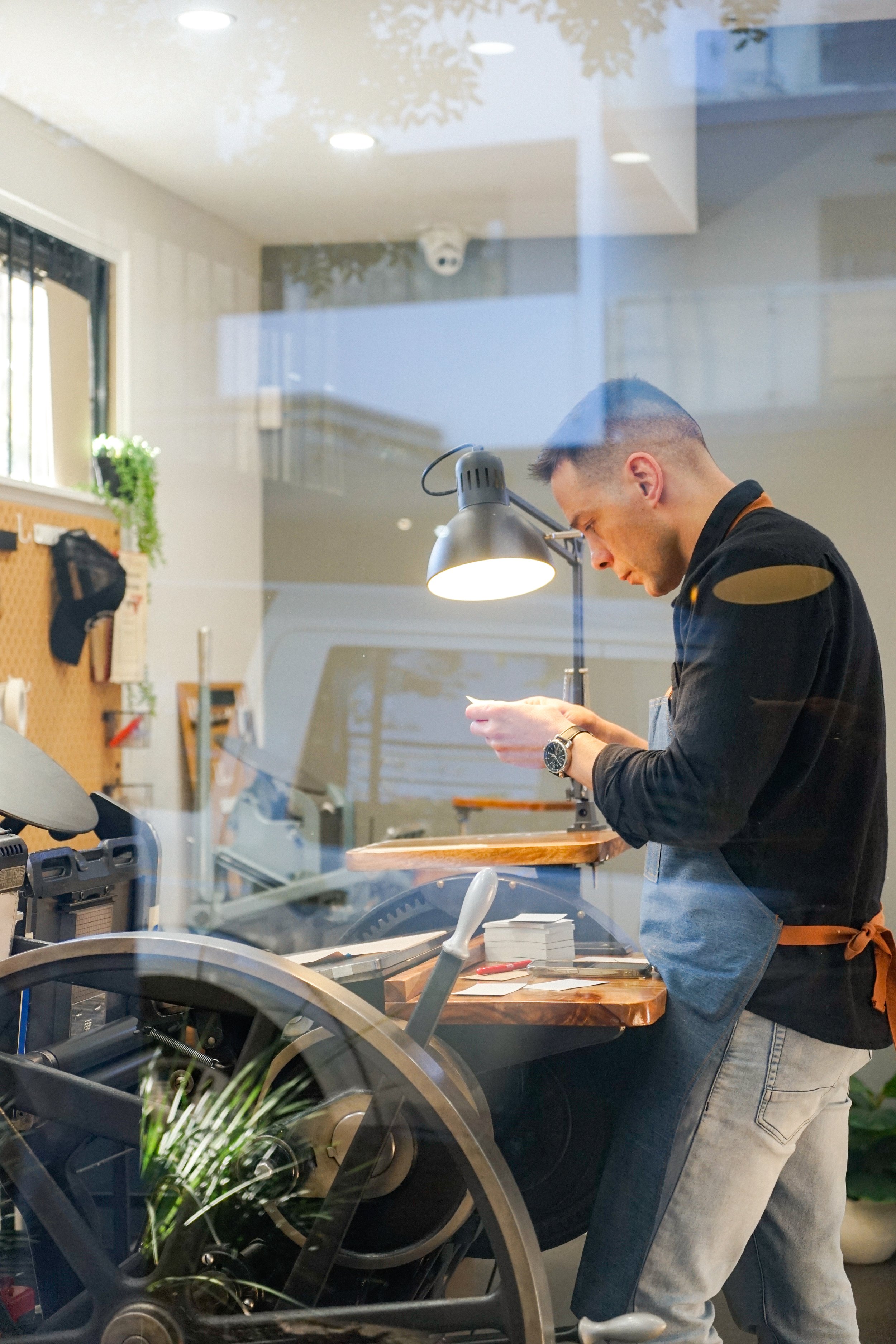 A man in a black shirt and jeans is standing at a work counter, looking down at his phone. The photo is taken through a window, showing reflective glass with some indoor lighting and a ceiling camera. The background has a pegboard with hanging tools and a small plant on the wall.