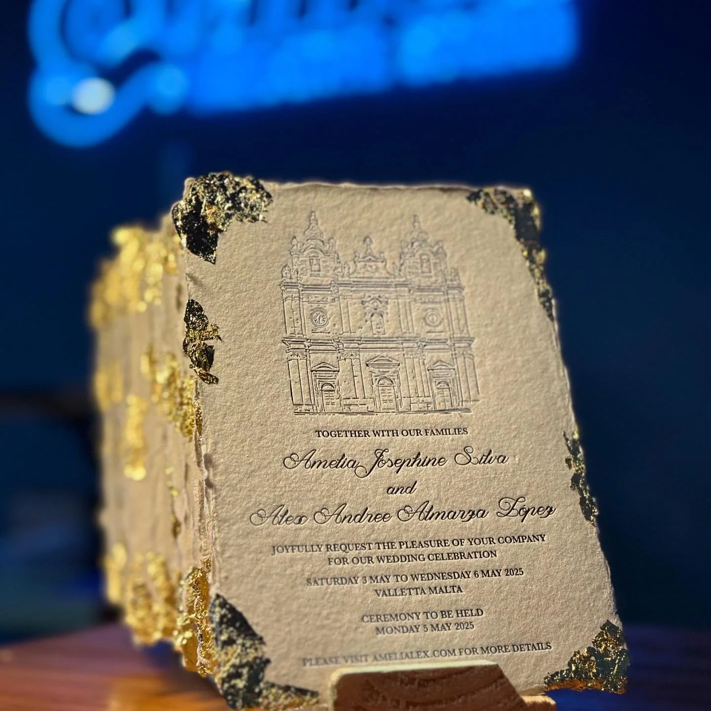 A wedding invitation with gold foil edges and an embossed illustration of a church, detailing the names of the couple and wedding details, set against a dark background.