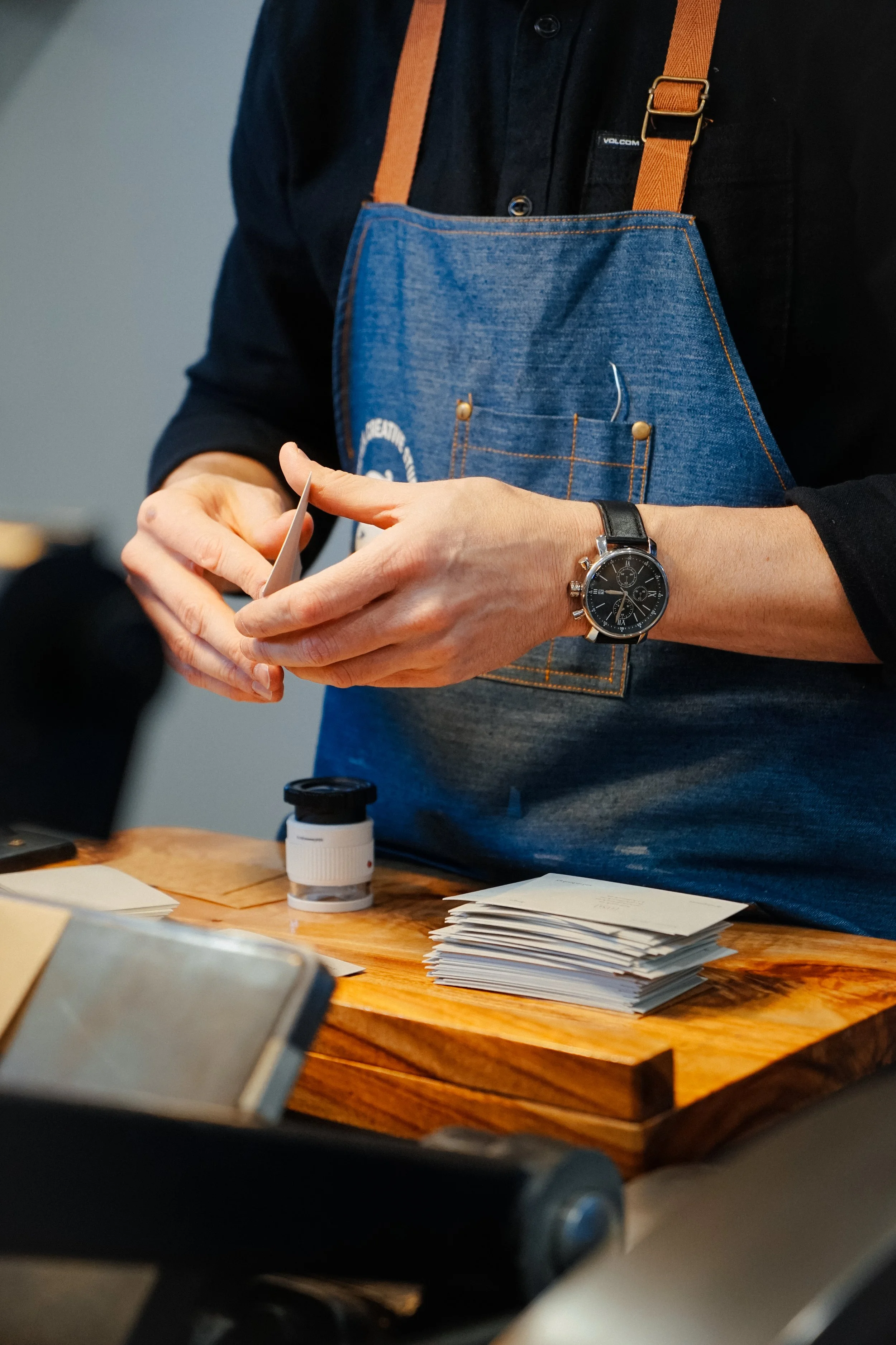 A person dressed in a black shirt and wearing a watch on their left wrist is holding a small slip of paper. They are standing next to a wooden table that has a pile of white papers, some bottles, and other objects on it. The person is also wearing a denim apron with orange straps, and a small device is attached to their apron pocket.
