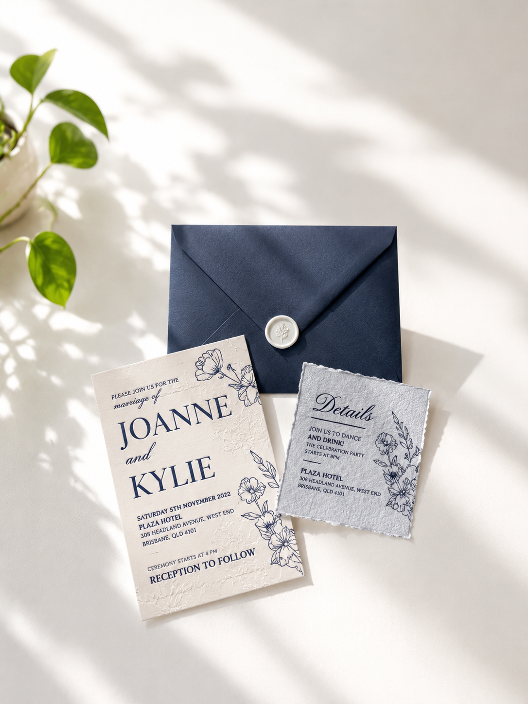 Wedding invitation card, details card, and navy envelope with a wax seal, on a white surface with shadows and a green plant in the background.
