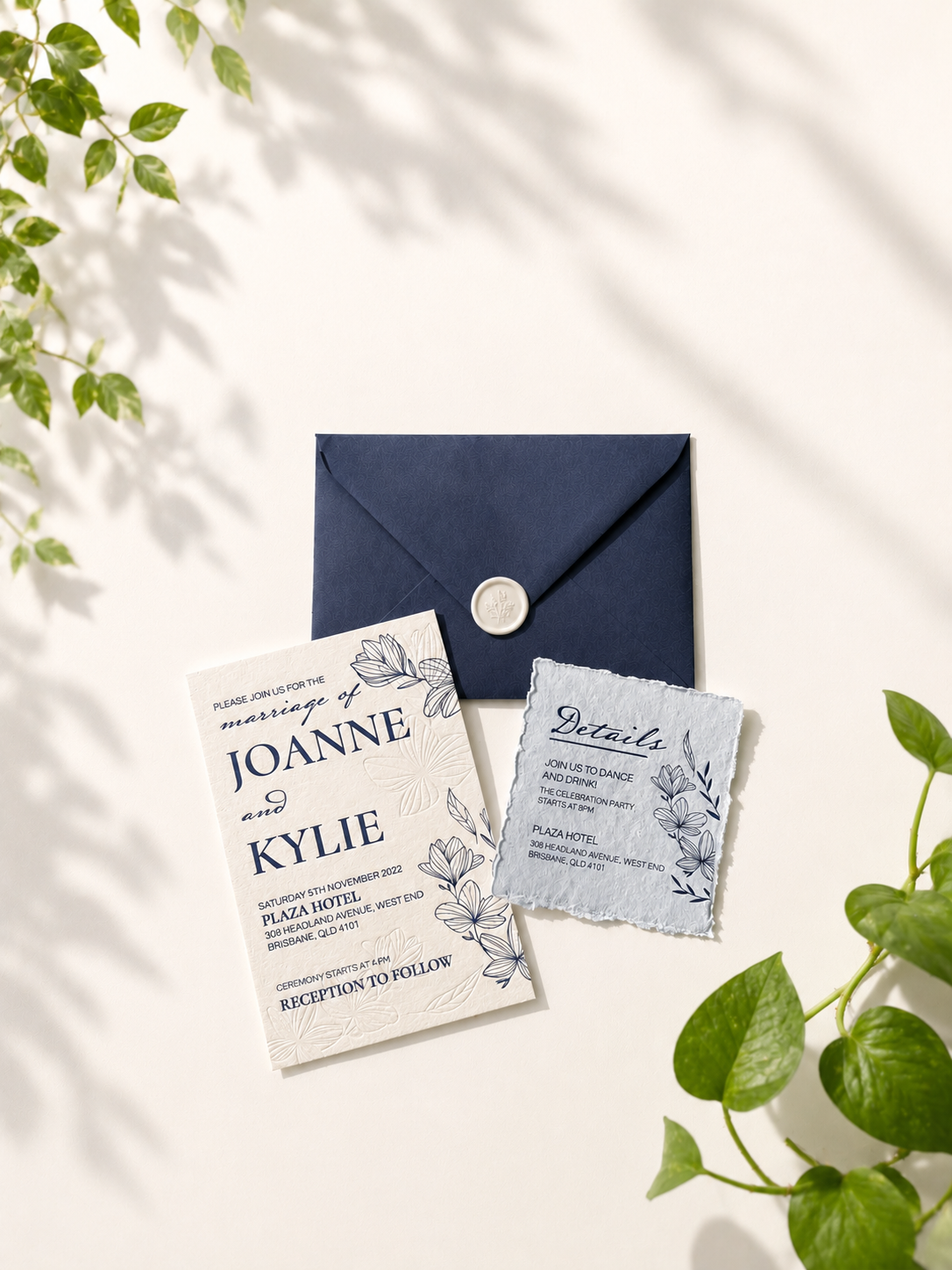 Wedding invitation card and details card with floral design, placed on a white surface beside a blue envelope and surrounded by green leaves.