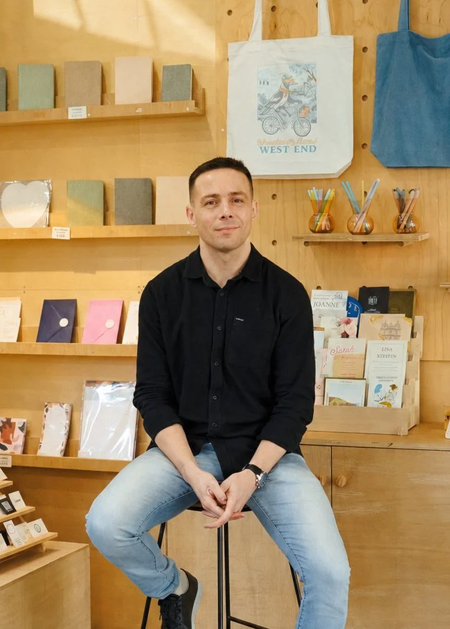 A man in a black shirt and light blue jeans sitting on a stool in a store with wooden walls, surrounded by notebooks, greeting cards, and colorful pens.