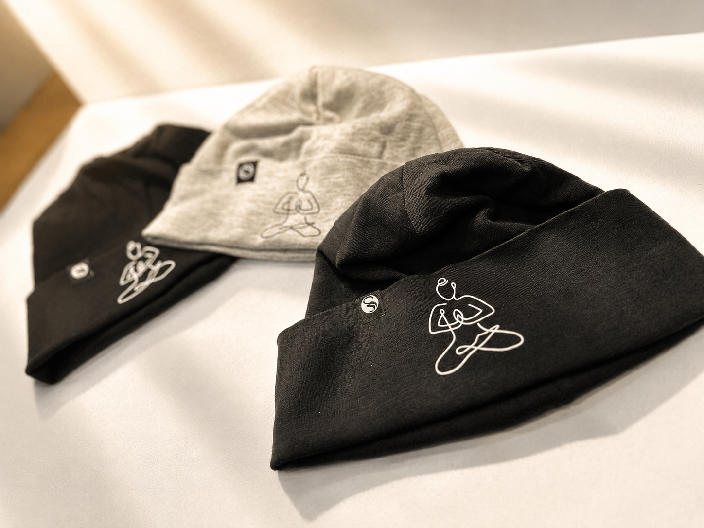 Three beanies in black, gray, and dark gray with minimalist line art of a person in a yoga pose embroidered on each, laid out on a white surface.