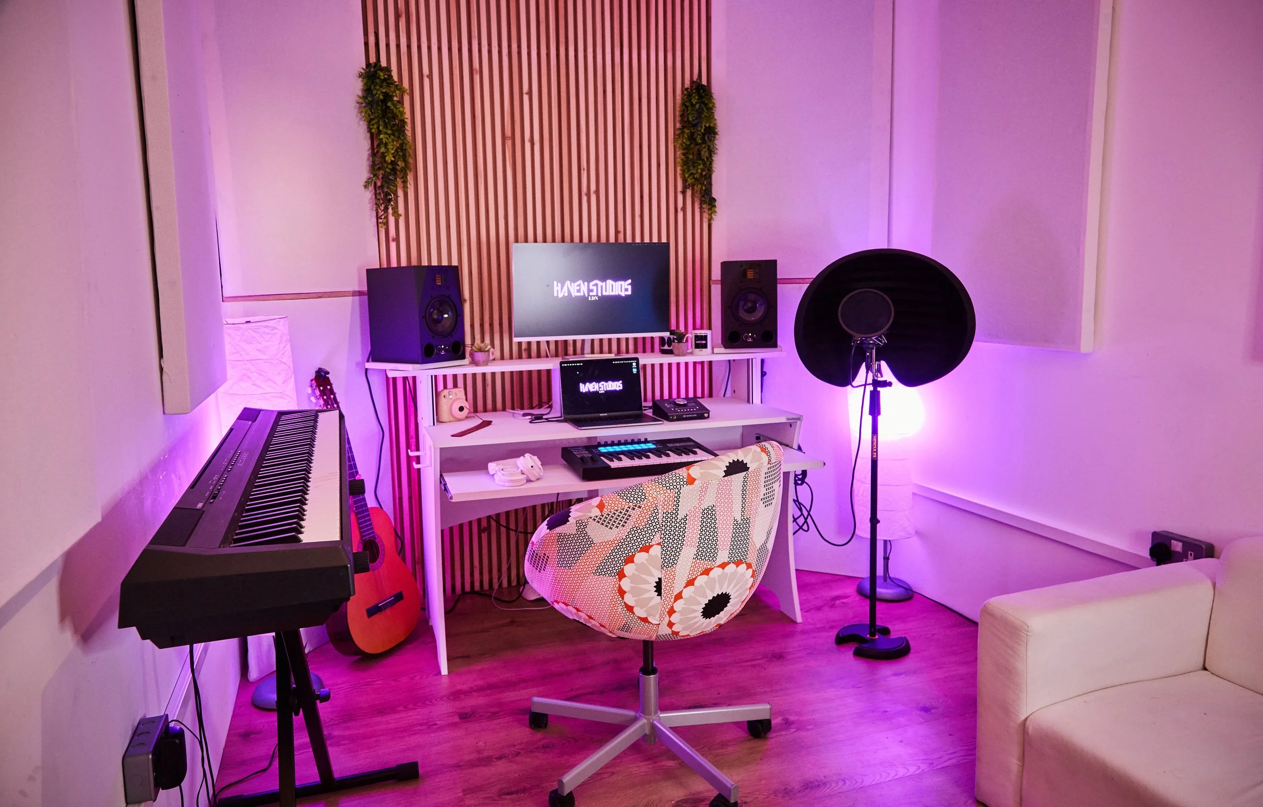 A music studio with a pink purple ambient light. There is a digital keyboard on a stand on the left, an acoustic guitar leaning against the wall, a desk with two speakers, a laptop, a small digital device, and a smoothie cup. In front of the desk is a colorful, patterned swivel chair. To the right is a microphone with a pop filter on a stand, and a cream-colored sofa on the right side.