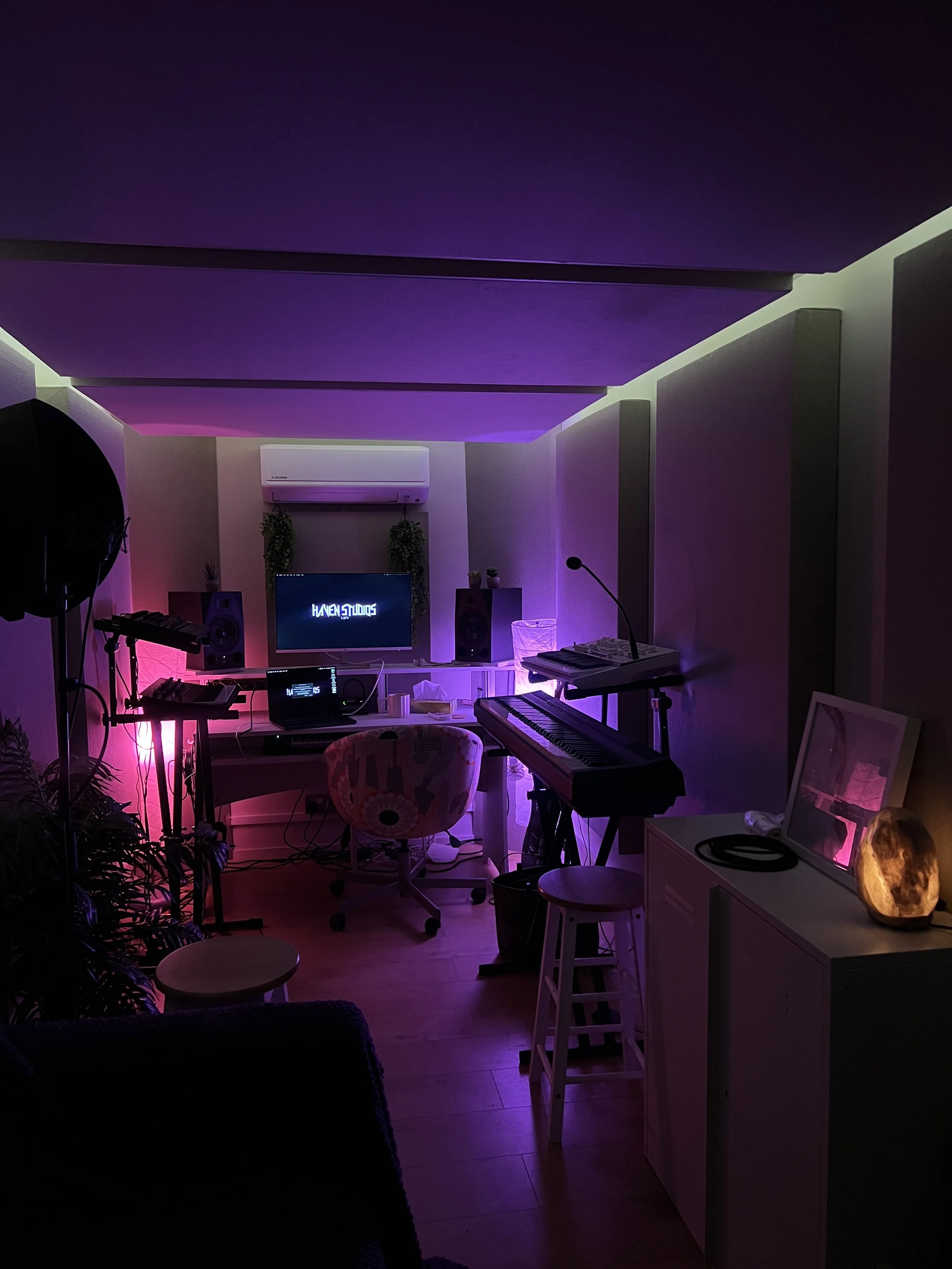 A music studio with purple ambient lighting, featuring keyboards, studio monitors, a computer monitor with the text 'Haven Studios', a microphone, and various musical equipment and decor.