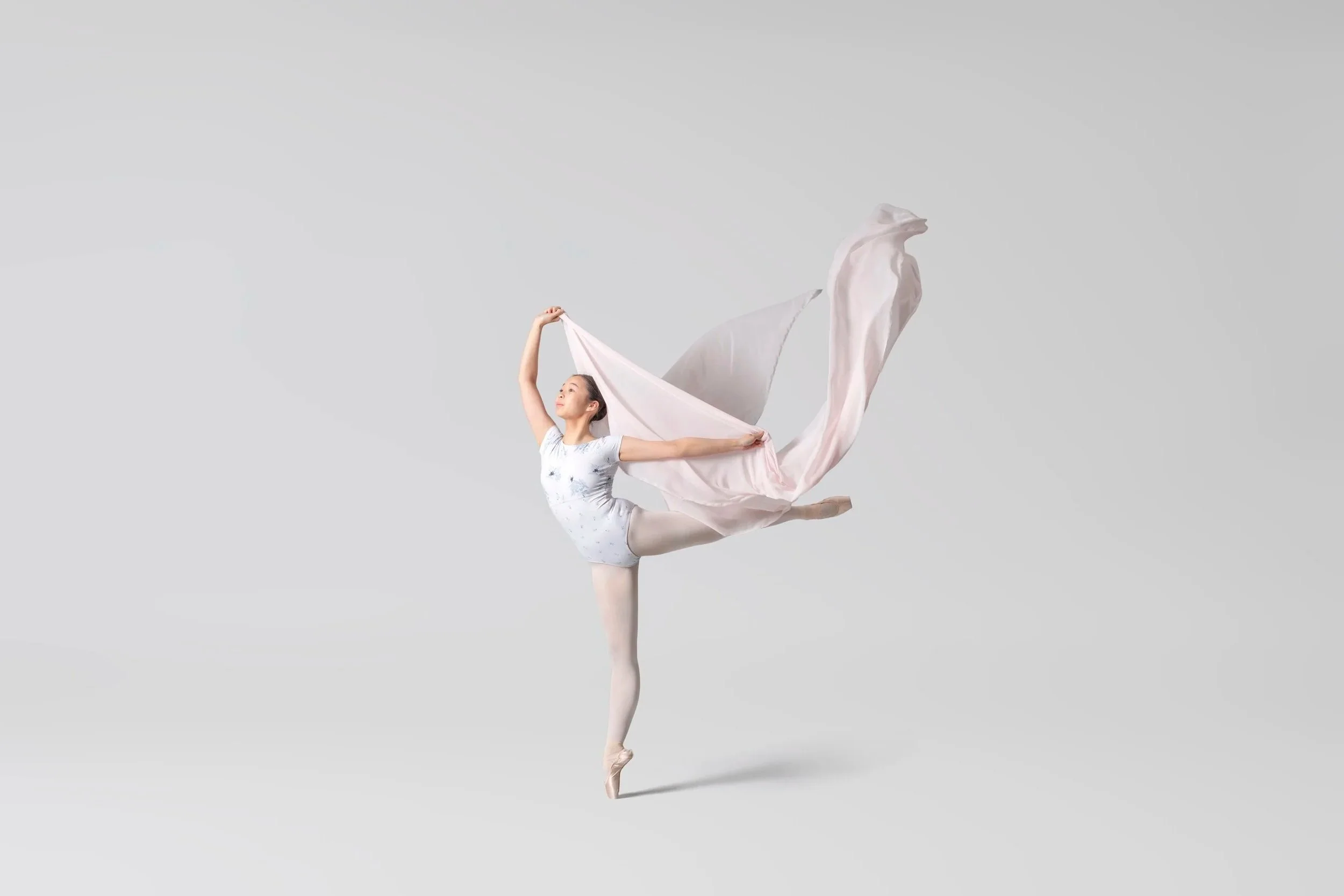 A ballet dancer in a white leotard and pink tutu performing a balanced pose on one leg with arms extended, holding a flowing pink fabric, against a plain gray background.
