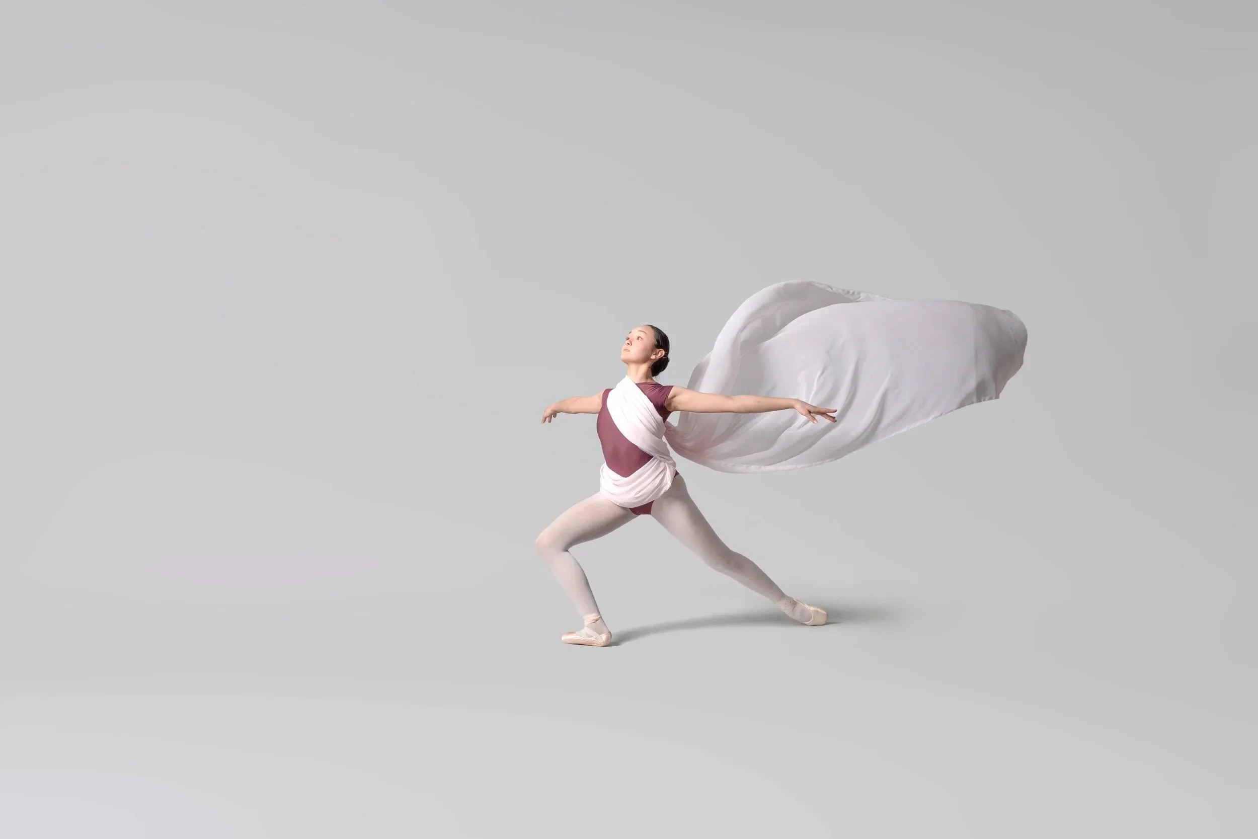 A ballet dancer wearing a pink and white costume, posed in a graceful ballet stance with arms extended and one leg bent, against a plain gray background, with a flowing white fabric attached to her back.