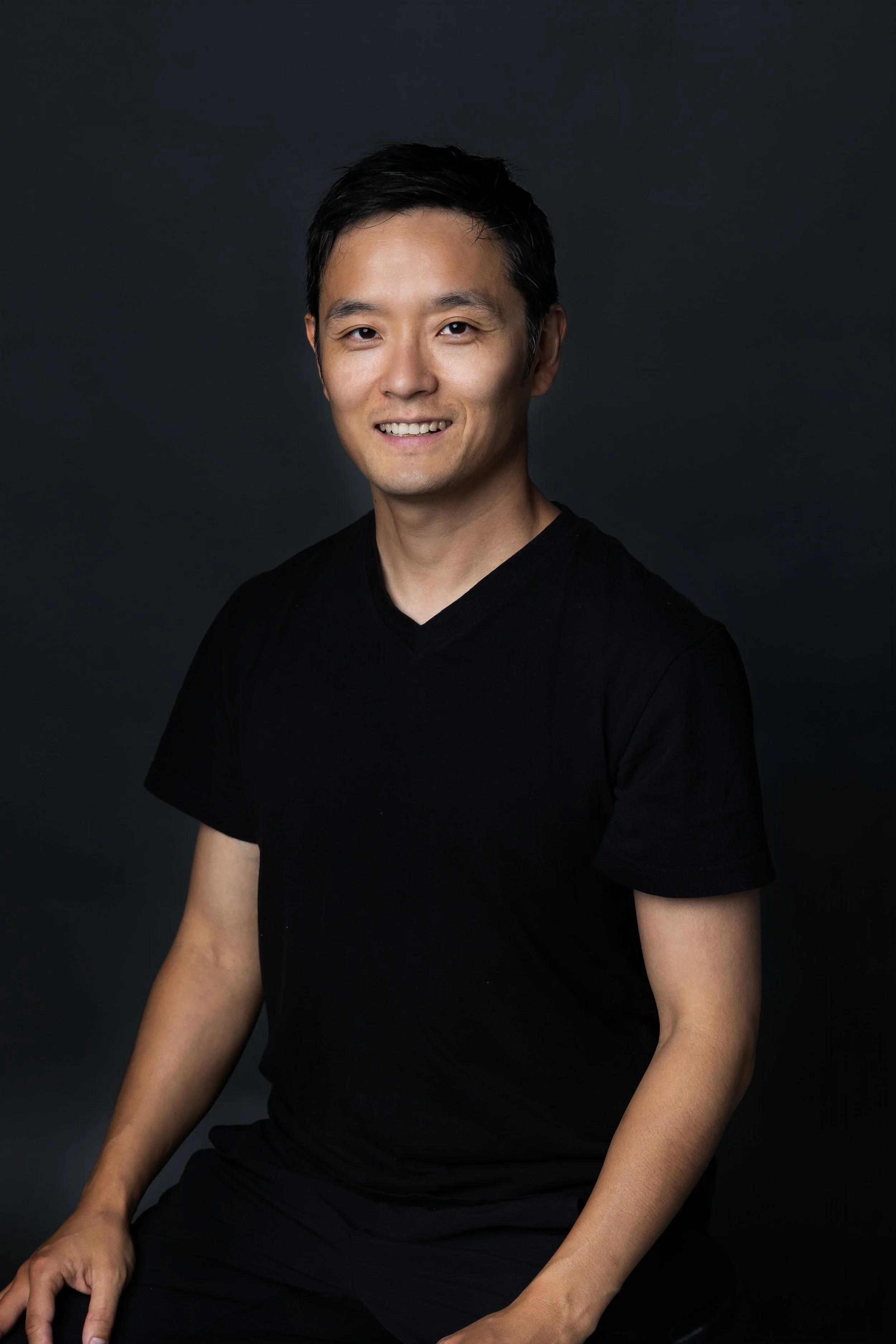 Portrait of a smiling Asian man in a black t-shirt against a dark background.