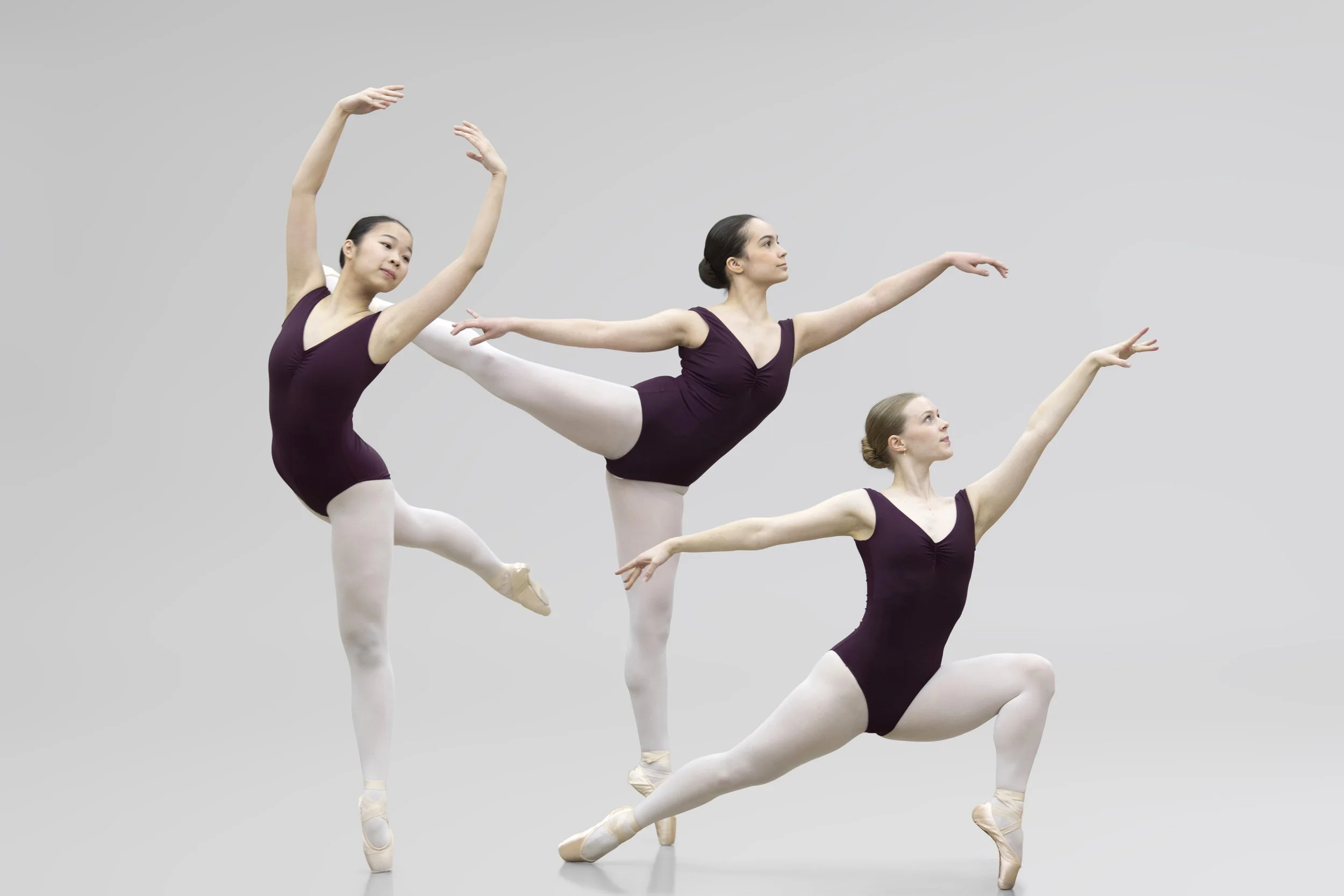 Three female ballet dancers in purple leotards and white tights performing ballet poses against a plain background.